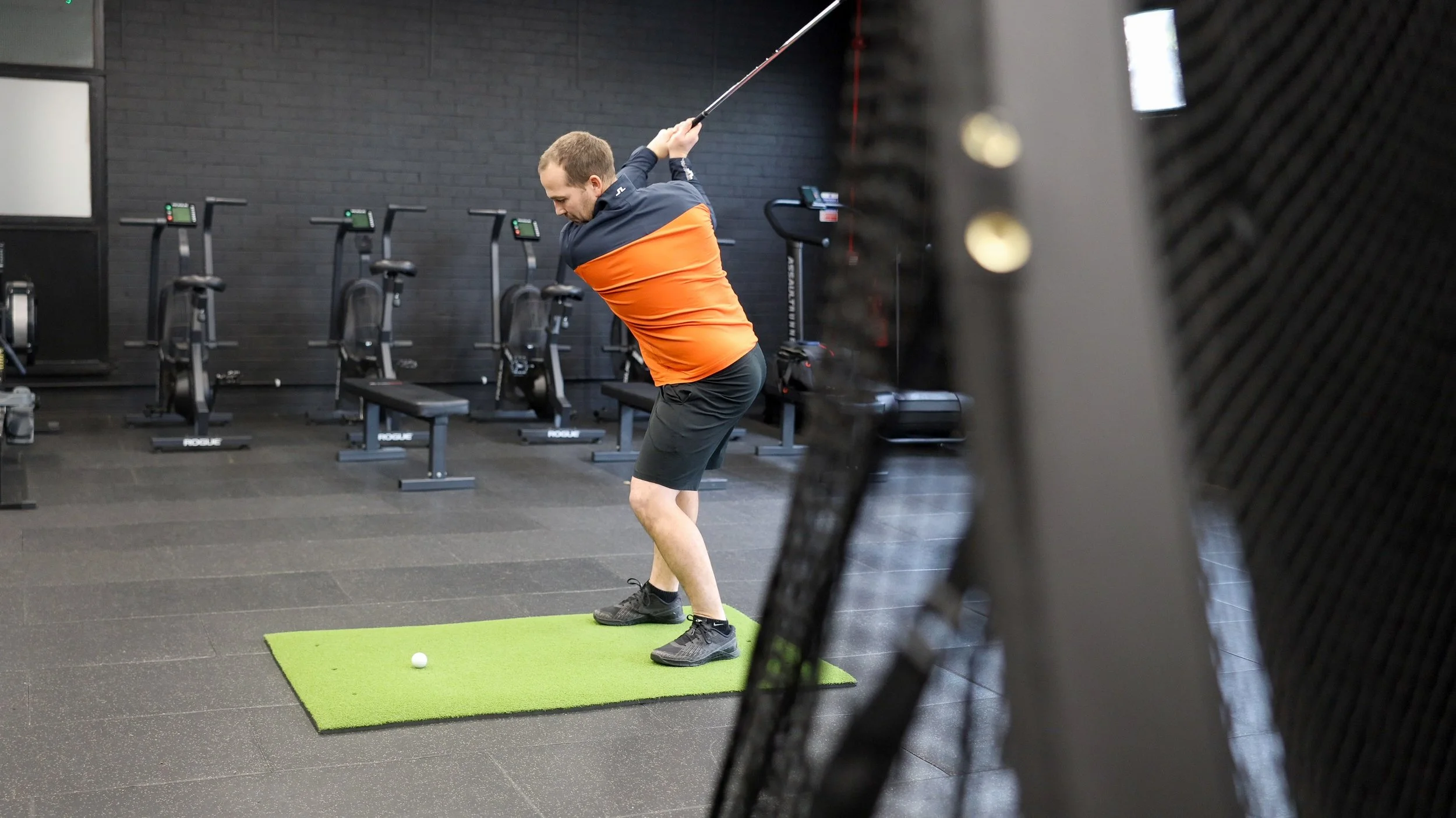 A man in a gym swings a golf club while standing on a green mat, with a golf ball in front of him. The gym has black brick walls, exercise bikes against the wall, and gym equipment visible in the background.