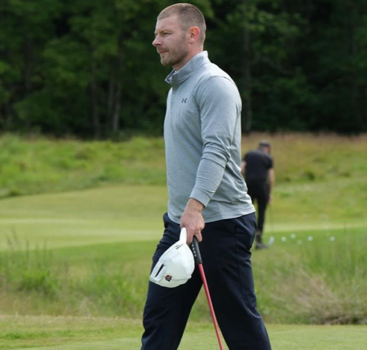 A man on a golf course holding a golf club and a golf ball marker, wearing a gray long sleeve shirt and navy pants, with trees in the background.