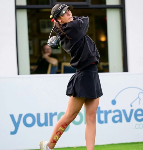 Young woman in black cap, black hoodie, and black skirt swinging a golf club at a driving range.