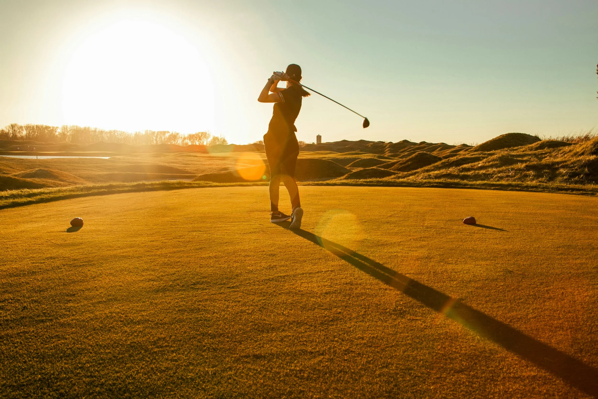 A person playing golf on a course during sunset, taking a shot with a club, with a bright sun low in the sky and landscape of grass and trees in the background.