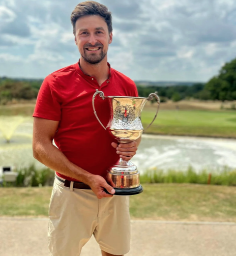 Smiling man in red shirt and beige shorts holding a large silver trophy on a golf course.