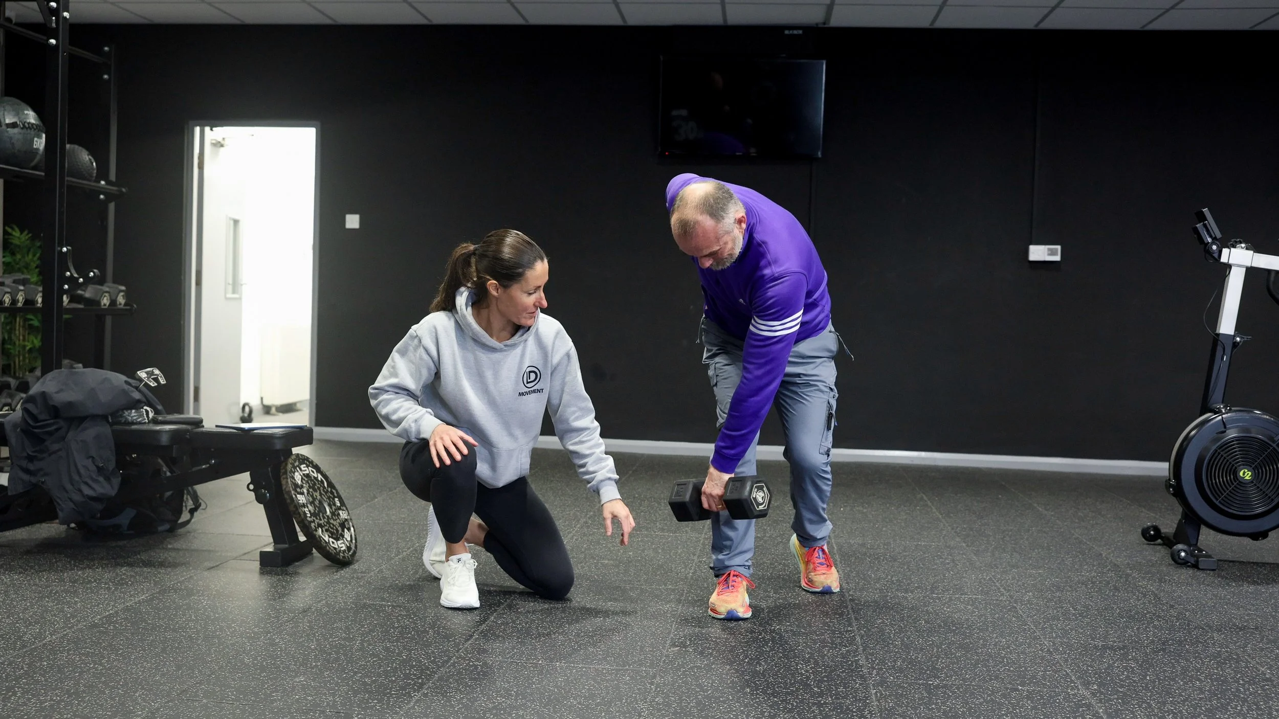 A woman and a man exercising together in a gym. The woman, wearing a gray hoodie, is kneeling on one knee and pointing to a dumbbell. The man, dressed in a purple long-sleeve shirt and gray pants, is holding a dumbbell and bending forward. There is gym equipment and a black wall in the background.