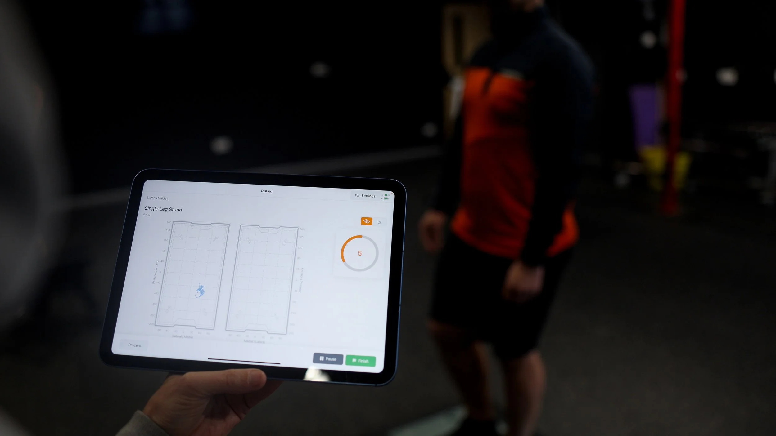 Tablet screen displaying a fitness app with a graph labeled 'Single Leg Stand' and a person in the background wearing a jacket, blurred out in a dark gym environment.