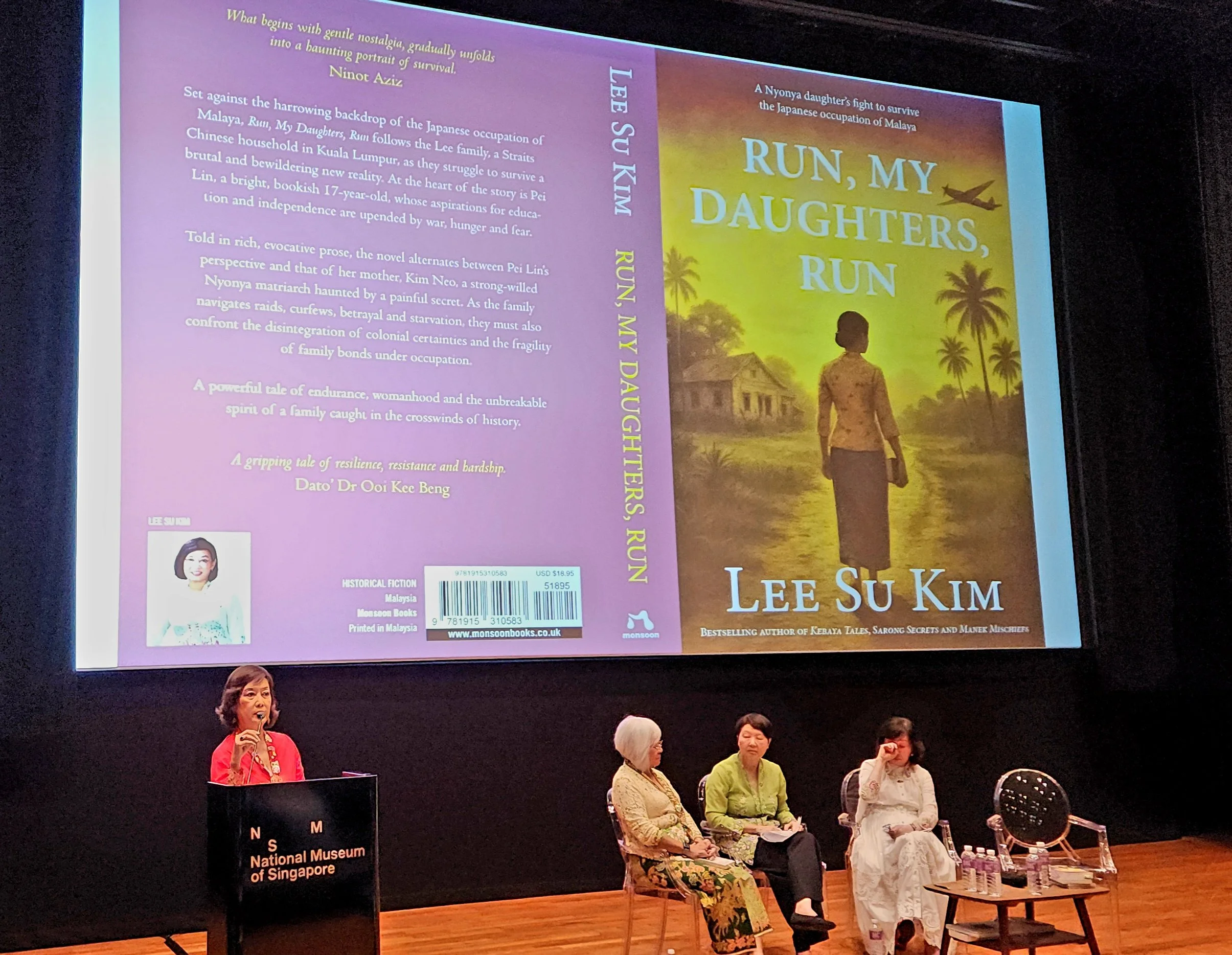 A group of people seated on stage during a panel discussion or presentation at the National Museum of Singapore, with a large book cover for 'Run, My Daughters, Run' by Lee Su Kim projected behind them. The book features an illustration of a woman walking through a rural area