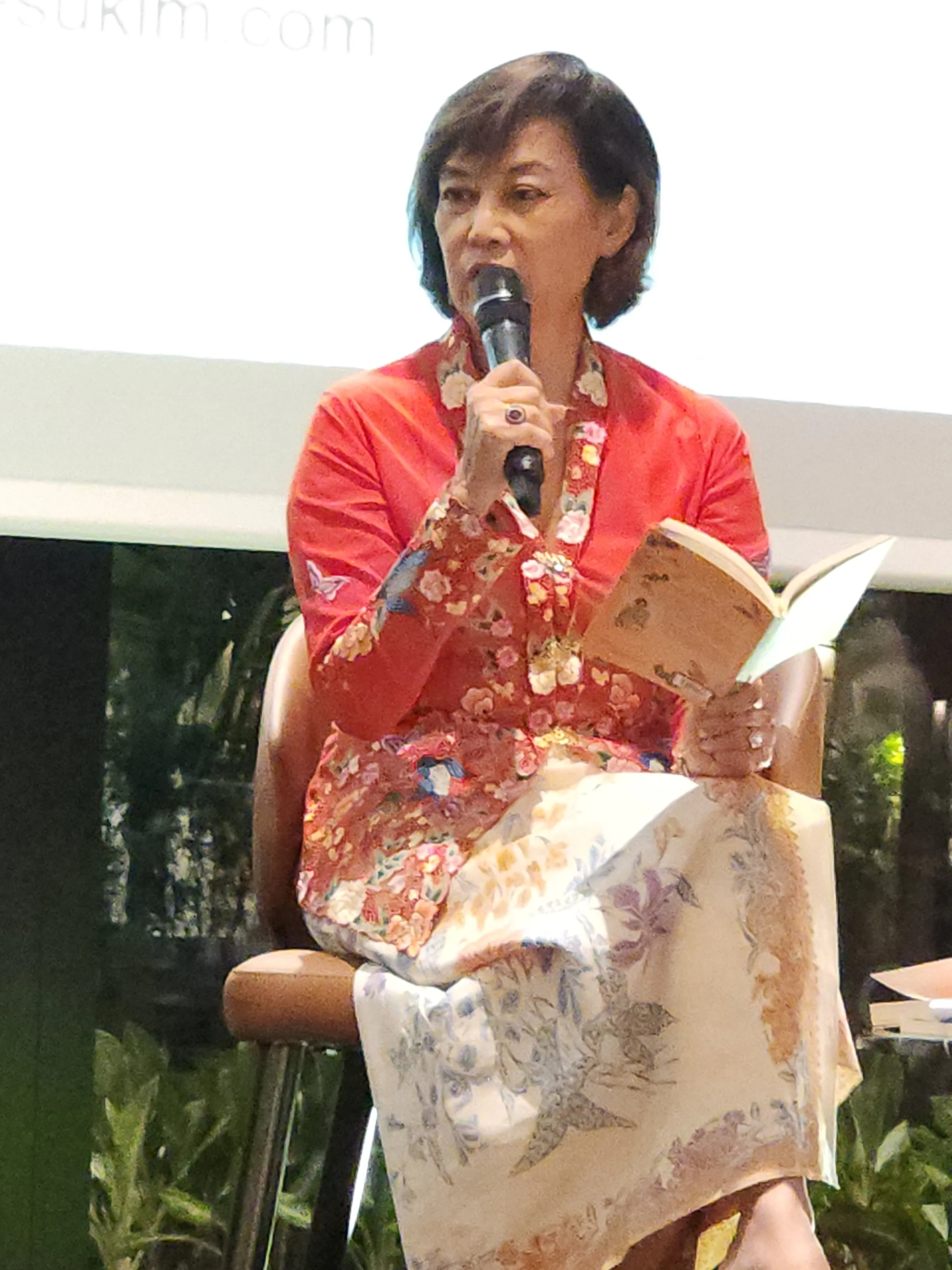 Woman wearing a red floral top reading from a piece of paper and speaking into a microphone.