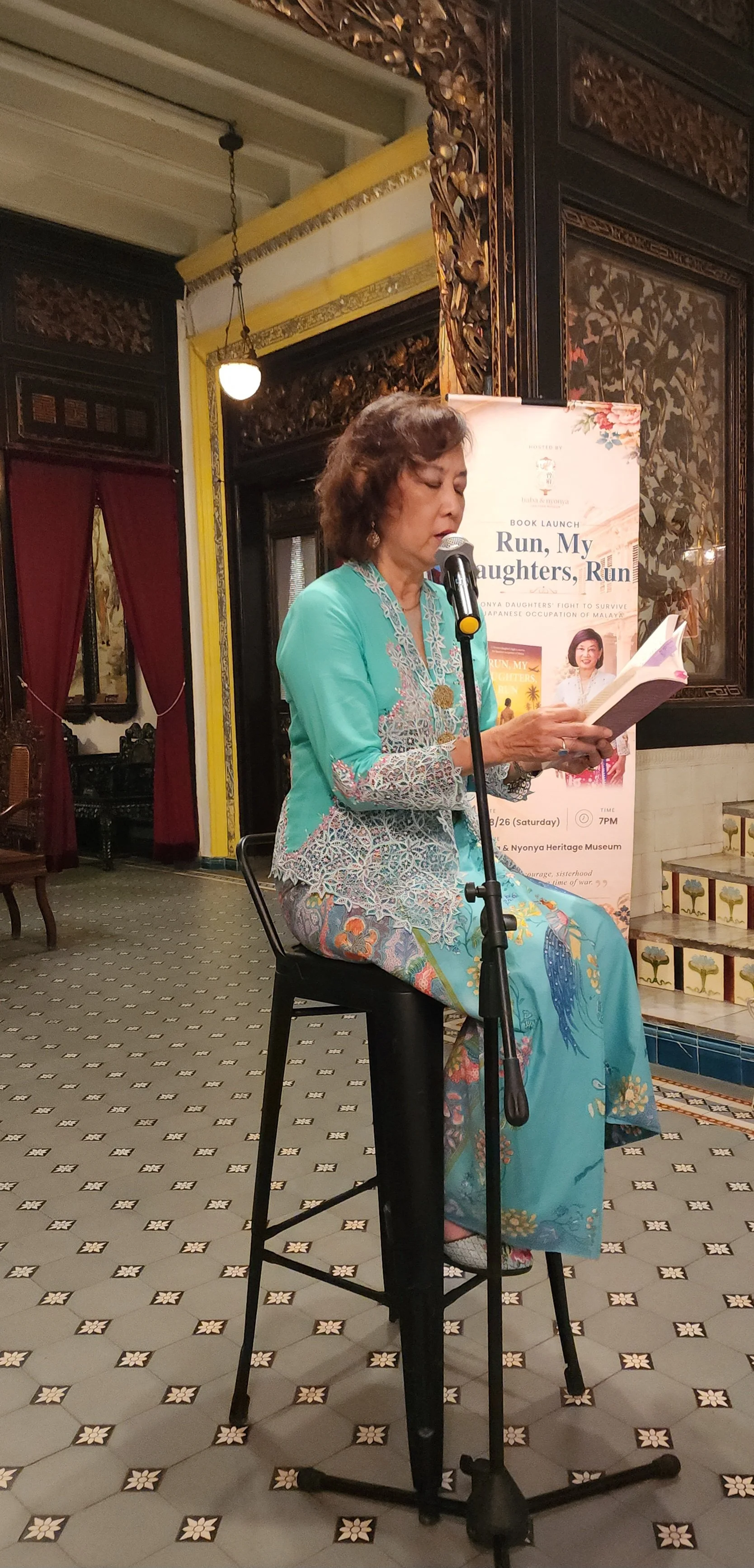 in traditional attire sitting on a black chair, reading from a book into a microphone at a book launch event. Behind her is a banner for the event, titled "Run, My Daughters, Run." The room has intricate wooden wall carvings, red curtains, and patterned tiled flooring.