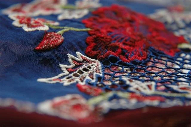 Close-up of embroidered fabric with red, white, and green thread designs on blue cloth. Sulam, a Nyonya style