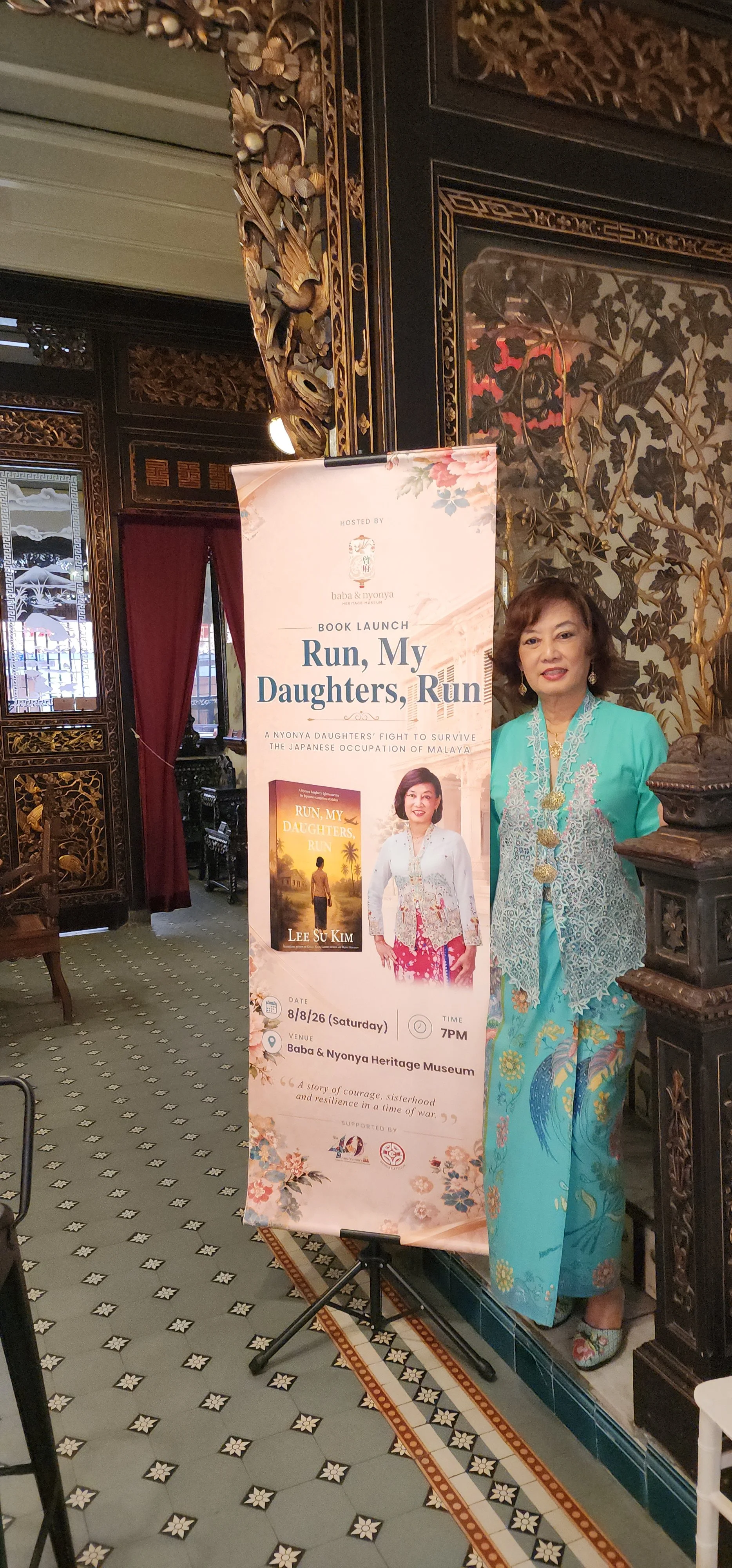 A woman in traditional attire standing next to a book launch poster for 'Run, My Daughters, Run' at the Baba & Nyonya Heritage Museum, with ornate wood and floral decor in the background.Lee su kim at her book launch