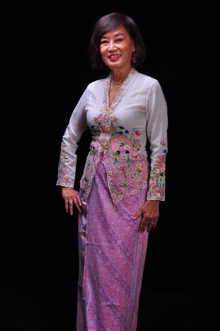 Lee Su KIm dressed in traditional Straits Chinese attire with intricate embroidery and a short-sleeved blouse kebaya, standing against a black background. Colourful Pekalongan batik sarong