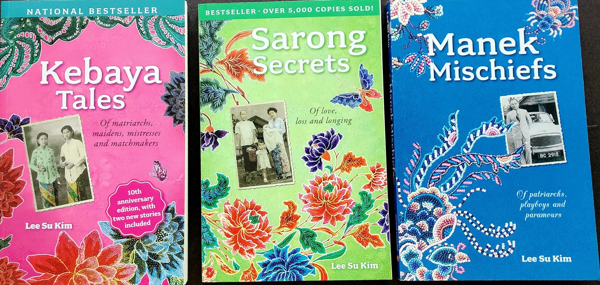 Three colorful books with floral and cultural designs on the covers. The pink book on the left is titled 'Kebaya Tales' and features black-and-white photographs of three women in traditional attire. The green book in the center is titled 'Sarong Secrets' and shows a black-and-white photo of a family, with decorative floral illustrations. The blue book on the right is titled 'Manek Mischiefs' and includes a black-and-white photo of a man in front of a car, with floral beadwork patterns.