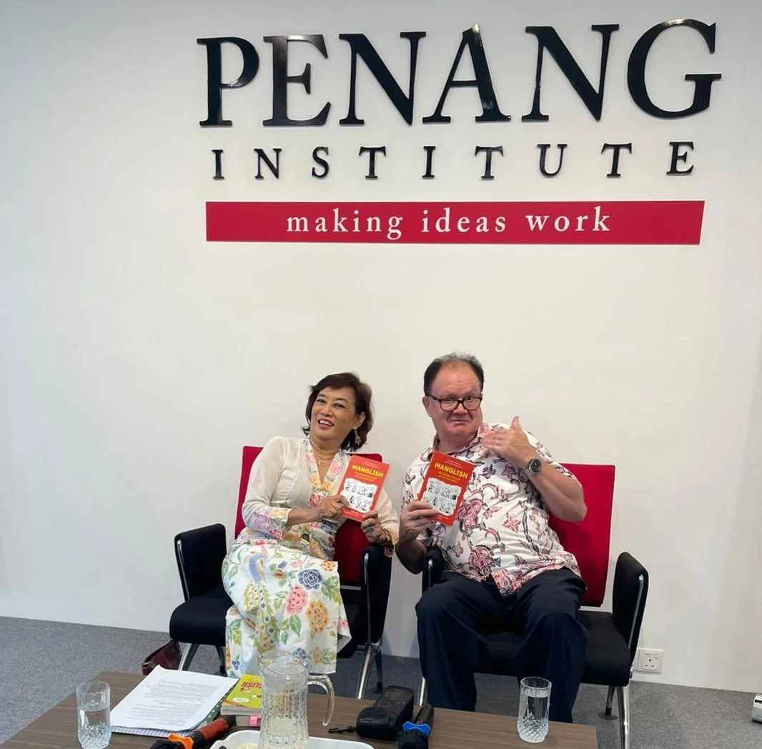 Two people sitting in chairs holding books at Penang Institute, with a wall sign reading 'making ideas work' behind them.