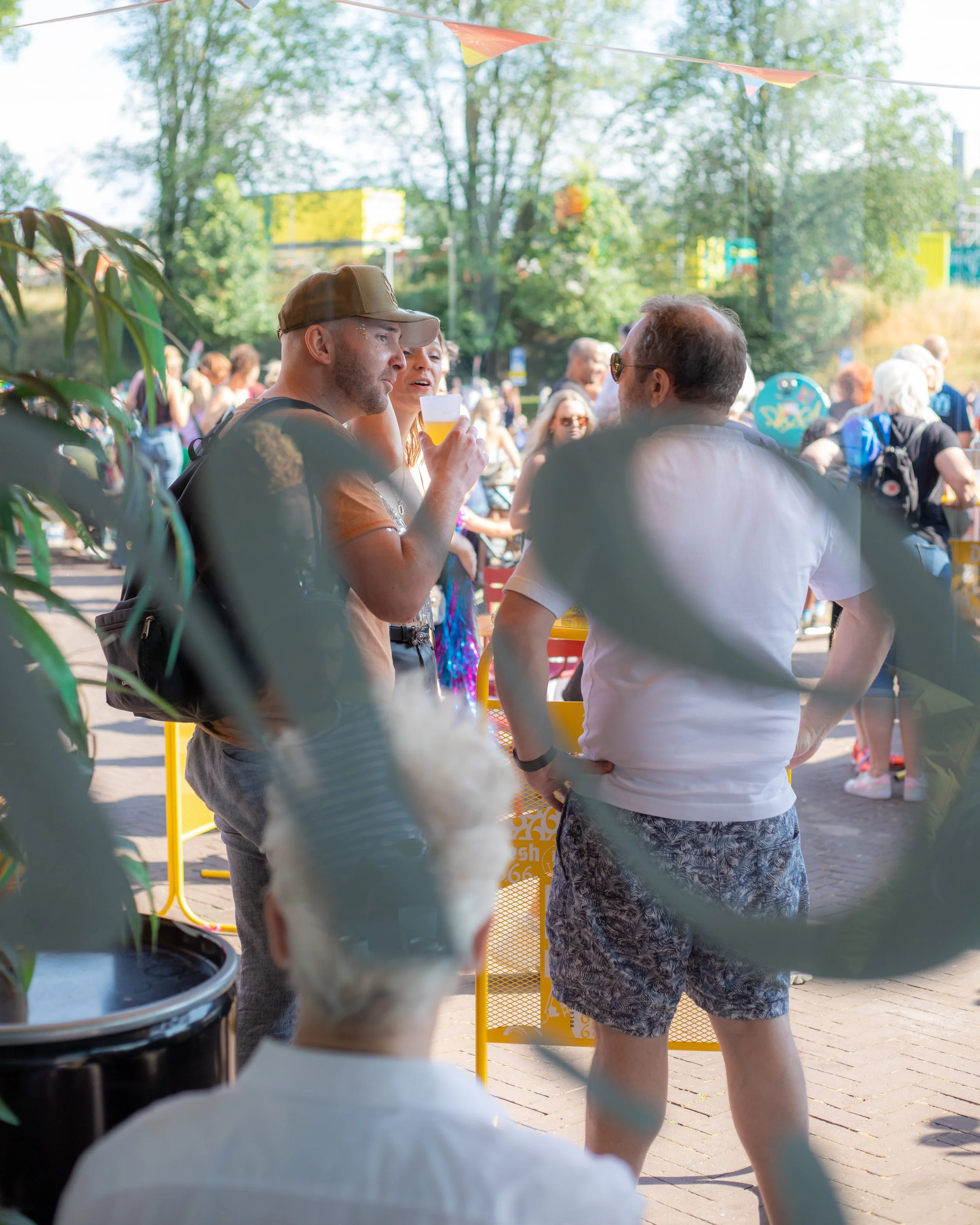 Mensen praten en drinken bier op een buitenevenement, gezien door een raampje met een bloempot op de voorgrond.