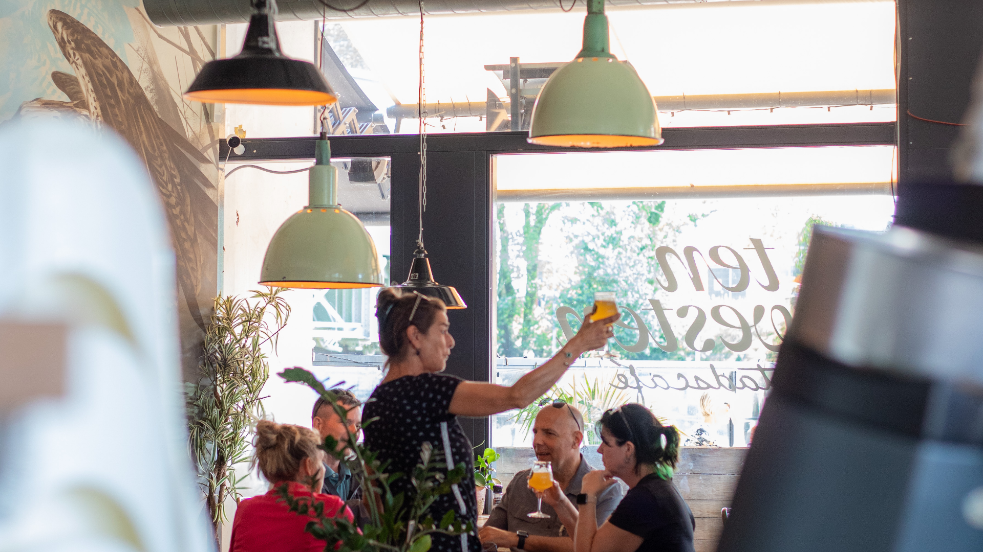 Binnen in een druk restaurant met groengeleide hanglampen, mensen die genieten en drinken, een serveerster uitreikend een glas, en decoratieve planten bij de ramen.