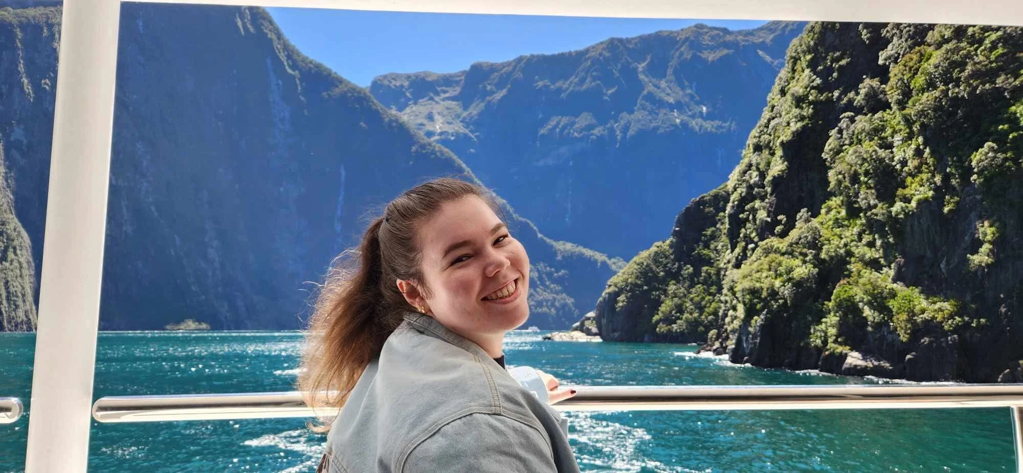 Tegan, young woman with pale skin and dark hair smiling at the camera. She is on a boat in Milford Sound, New Zealand with blue waters and a clear sky behind her, there are also mountains in the distance.