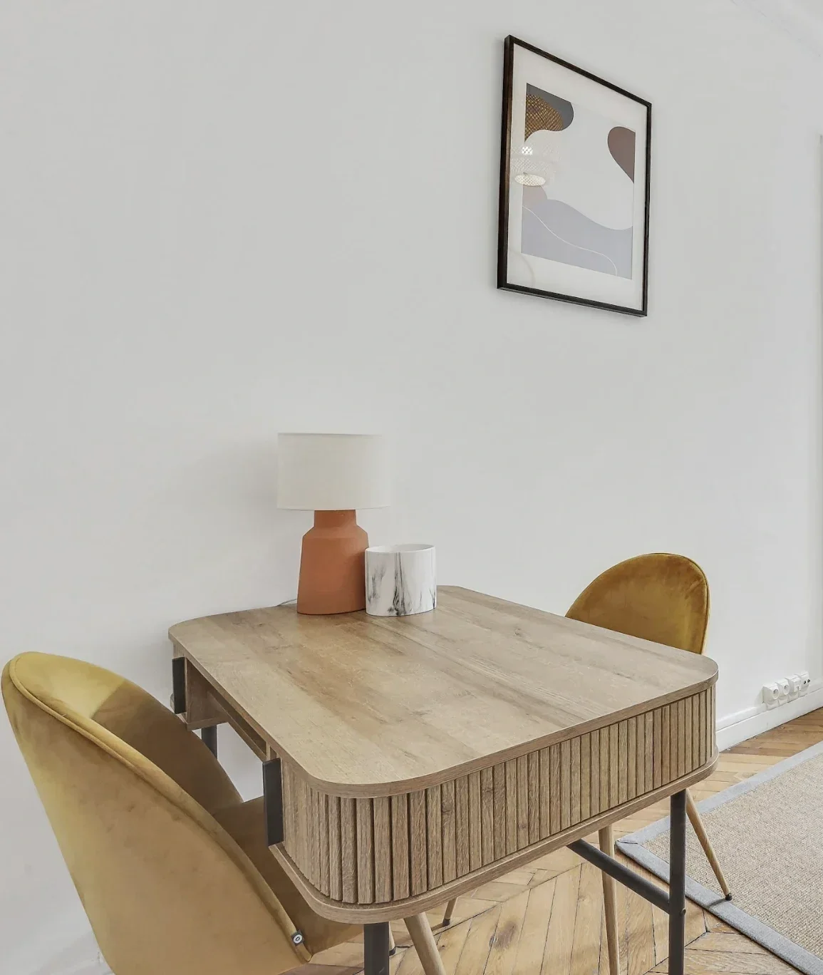 Une table en bois avec deux chaises en velours jaune, une lampe sur la table, un tableau abstrait accroché au mur blanc.