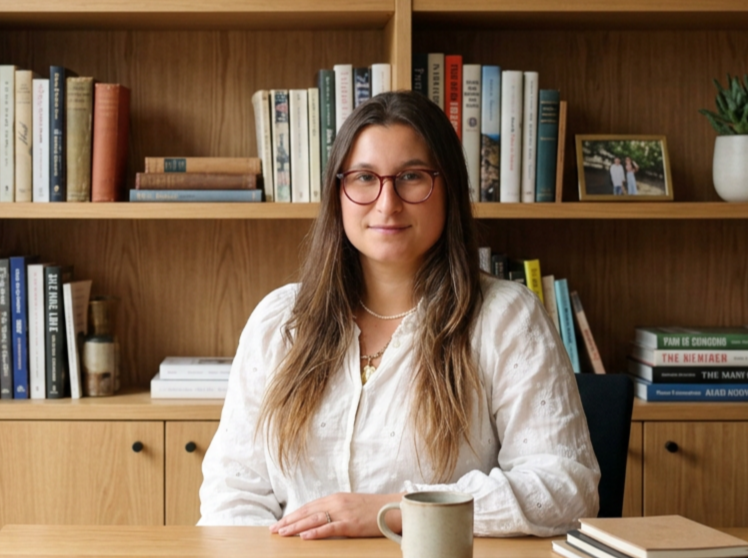 Femme assise à une table avec des livres en arrière-plan, portant des lunettes et un chemisier blanc, devant un mug sur la table.