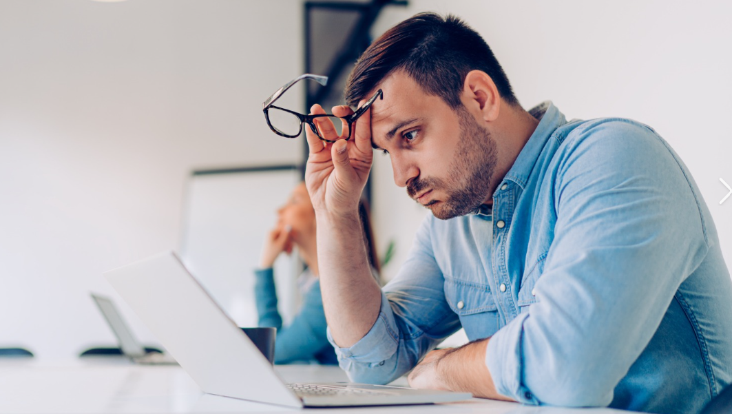 Man looking frustrated at work while holding glasses with computer and whiteboard in background