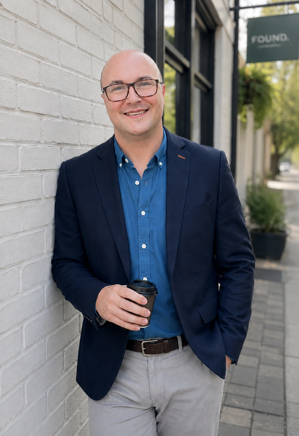 A smiling man with glasses and a bald head, wearing a navy blazer over a blue shirt, holding a coffee cup, standing outdoors next to a white brick wall.