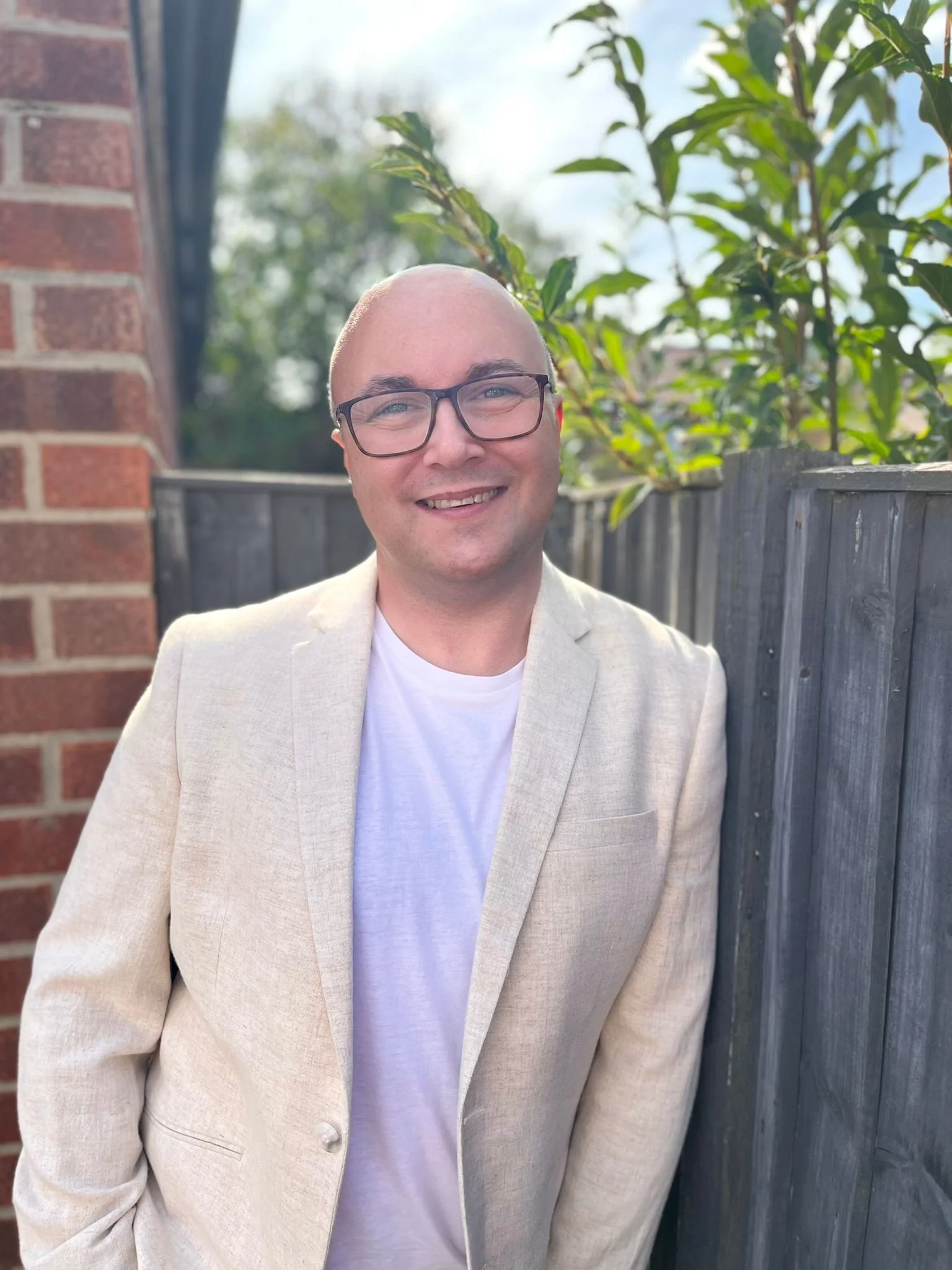 A man with a bald head and glasses, wearing a beige blazer and a white T-shirt, smiling outdoors next to a wooden fence and green plants.
