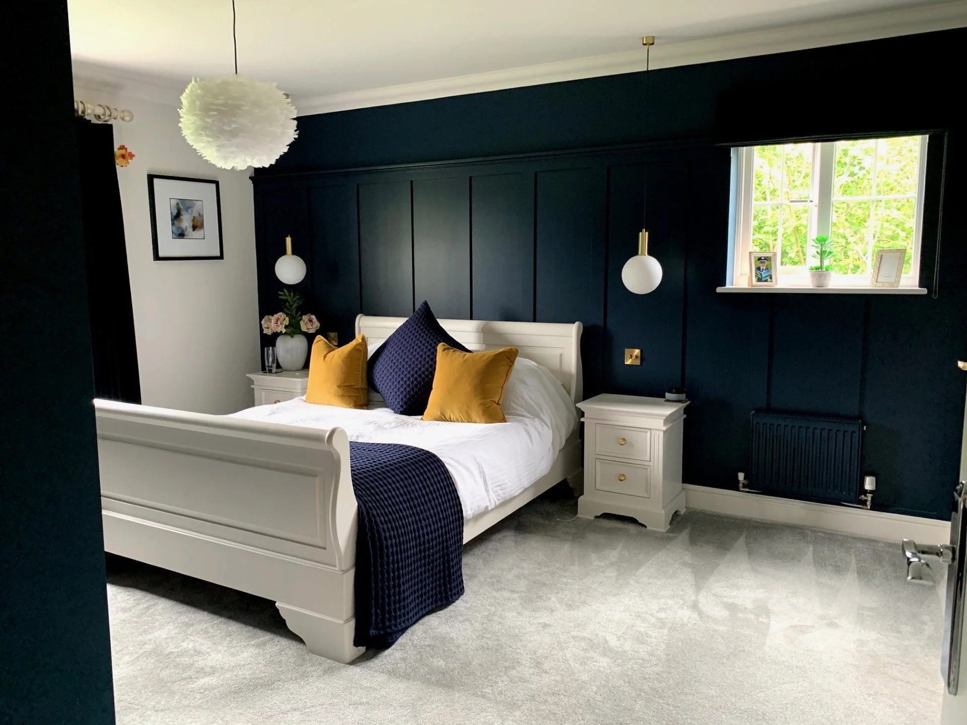 A bedroom with a white bed, navy blue and yellow pillows, and a navy blue bed cover. The room has dark blue wall panels, a window with a potted plant and family photos on the sill, and a nightstand with a vase of flowers on top. A modern light fixture hangs from the ceiling.