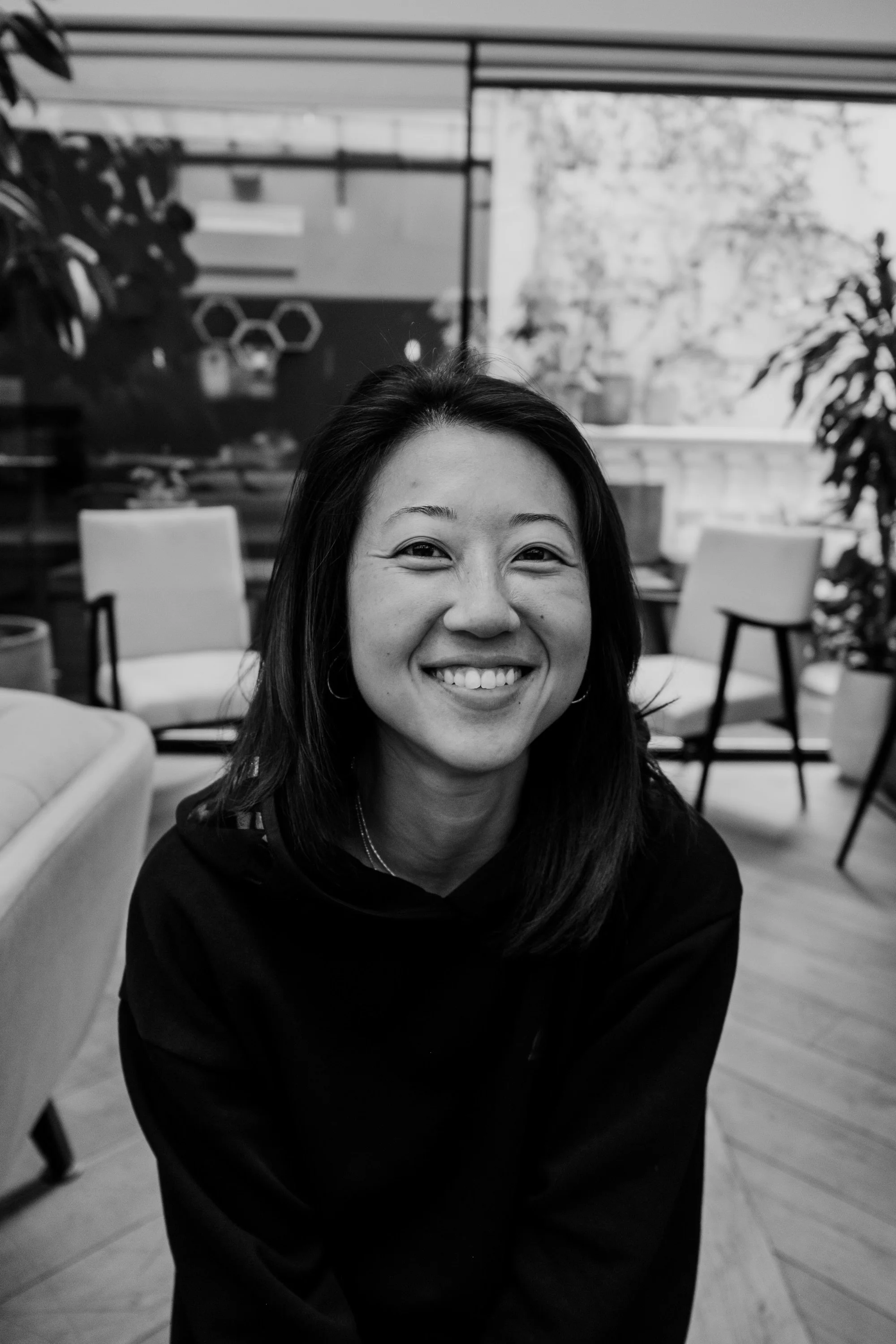 A woman with shoulder-length dark hair smiling in a black top in an indoor setting with chairs and plants in the background.