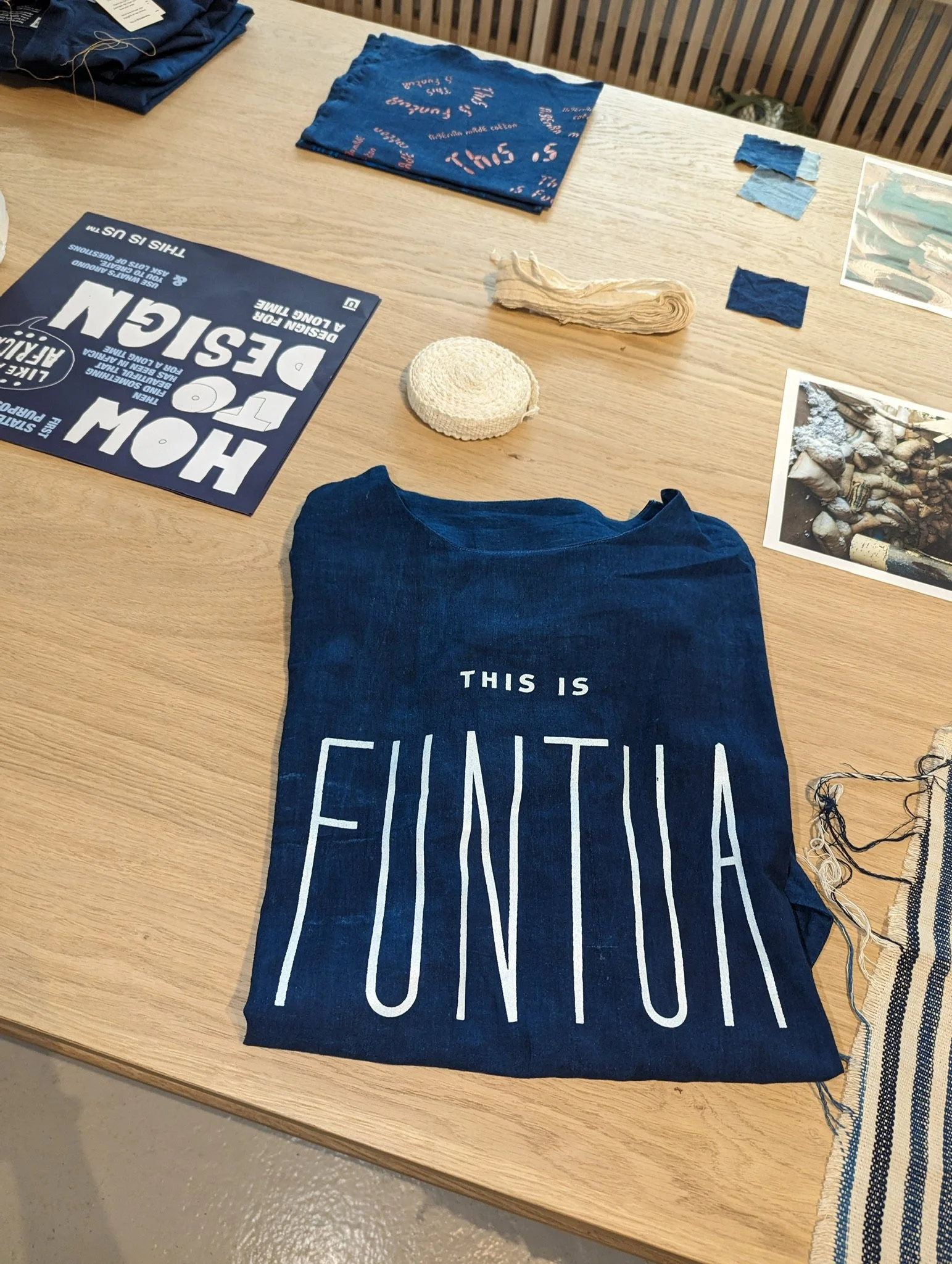 A wooden table with various items including folded blue fabric, a rolled beige fabric, a blue cloth with pink text, and a printed photograph of rocks and plastic debris. One blue fabric has text that reads "This is" and the other shows the answer, "C
