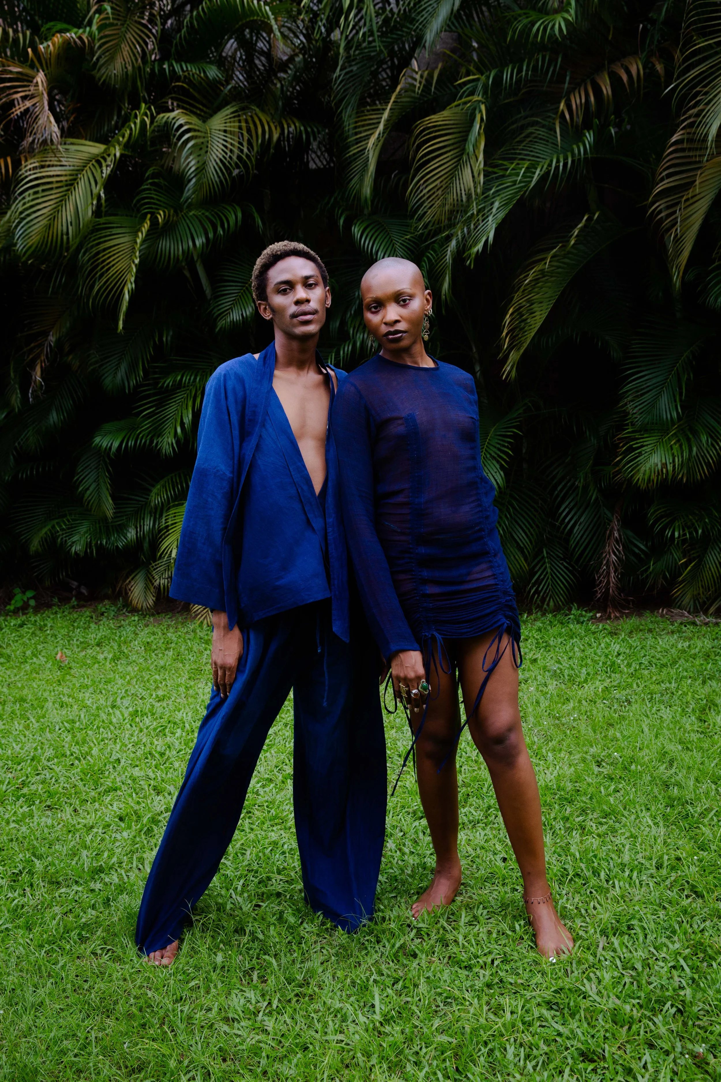 Two individuals stand on grass in front of lush green tropical plants, wearing fashionable blue clothing and striking confident poses.