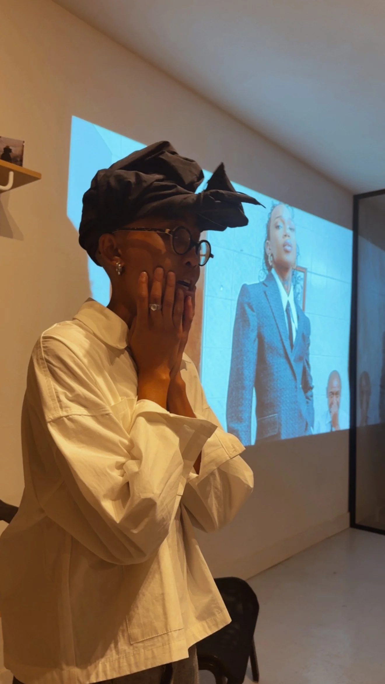 A woman wearing glasses and a head wrap, standing with her hands on her face, looking towards a screen displaying a woman in a suit.