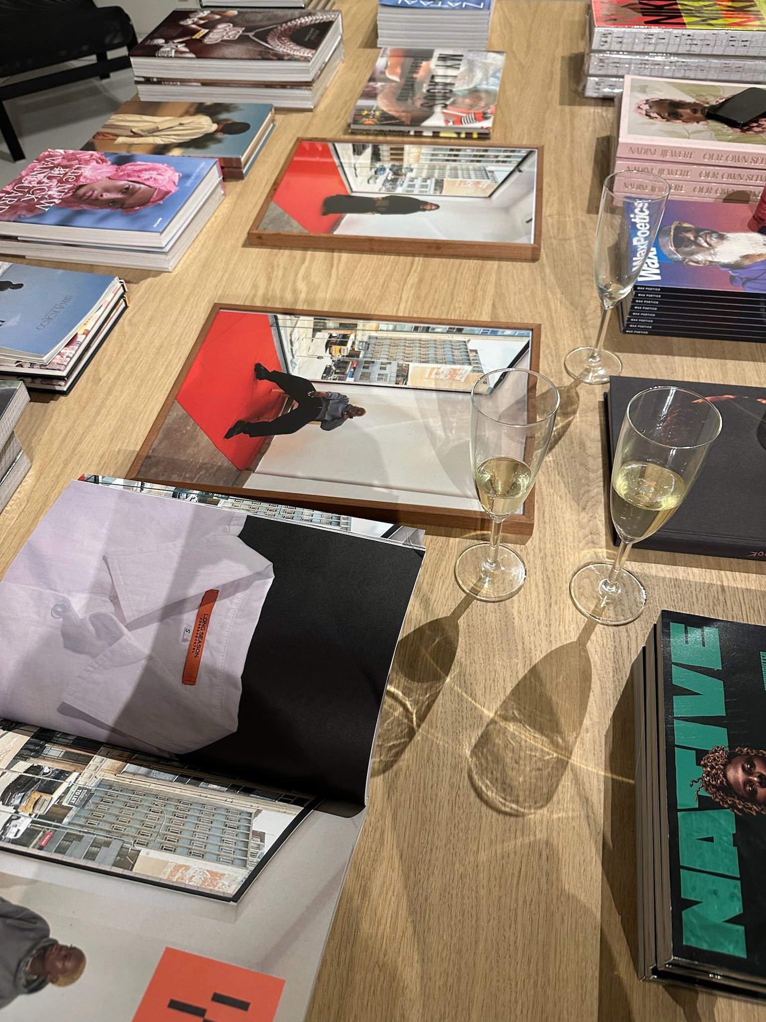 Books and magazines displayed on a wooden table, with three mirrors reflecting the room's interior, including a person standing on a red carpet.