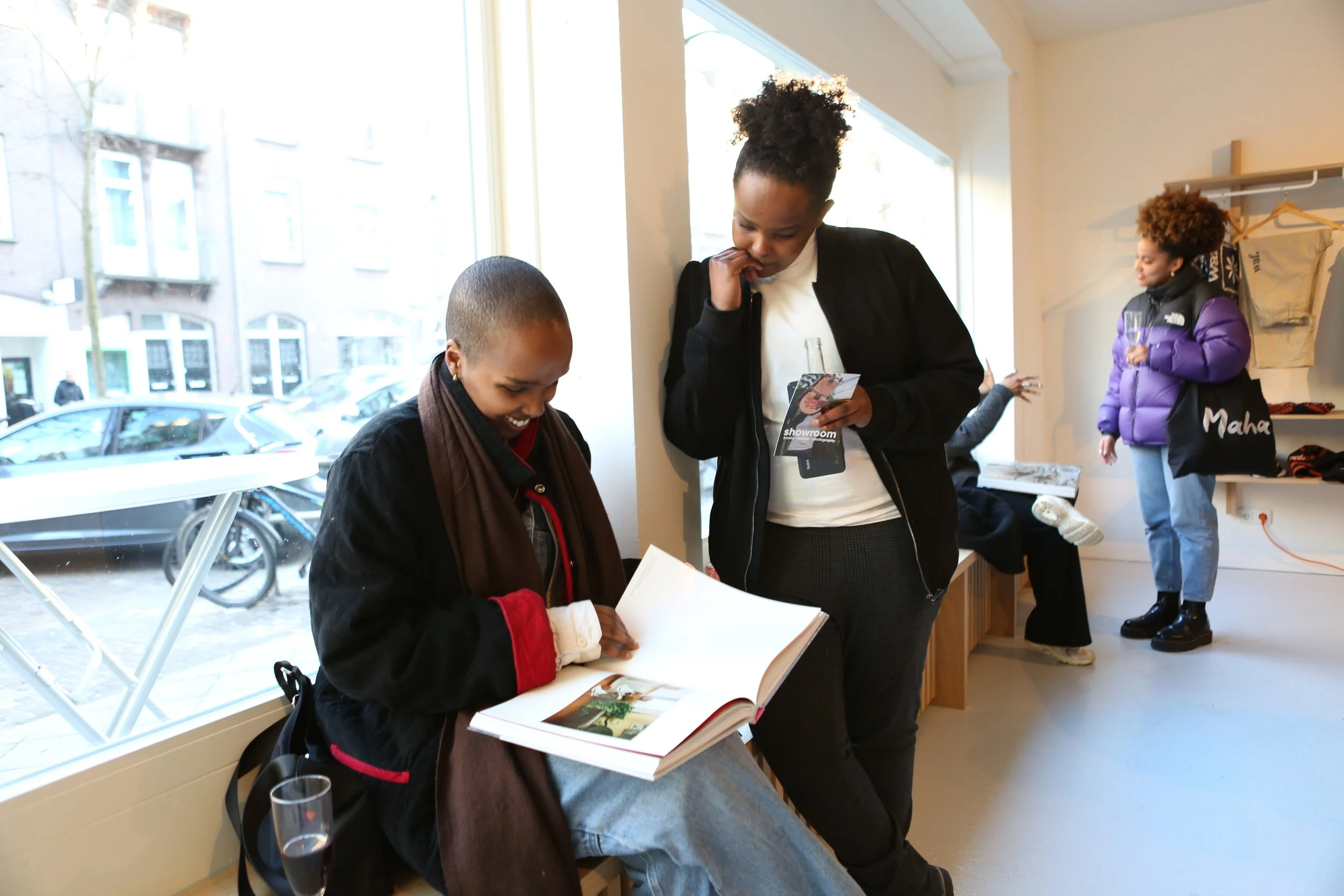 Two women are standing near a window inside a store or cafe, looking at a book or magazine. One woman has dark skin and curly hair tied up, wearing a black jacket, and holding a beverage in her hand. The other woman has lighter skin, short hair, and is wearing a black jacket with a scarf. A third woman with brown, curly hair, wearing a purple jacket, is standing in the background, holding a beverage and smiling. Outside the window, cars and a bicycle are parked on the street.