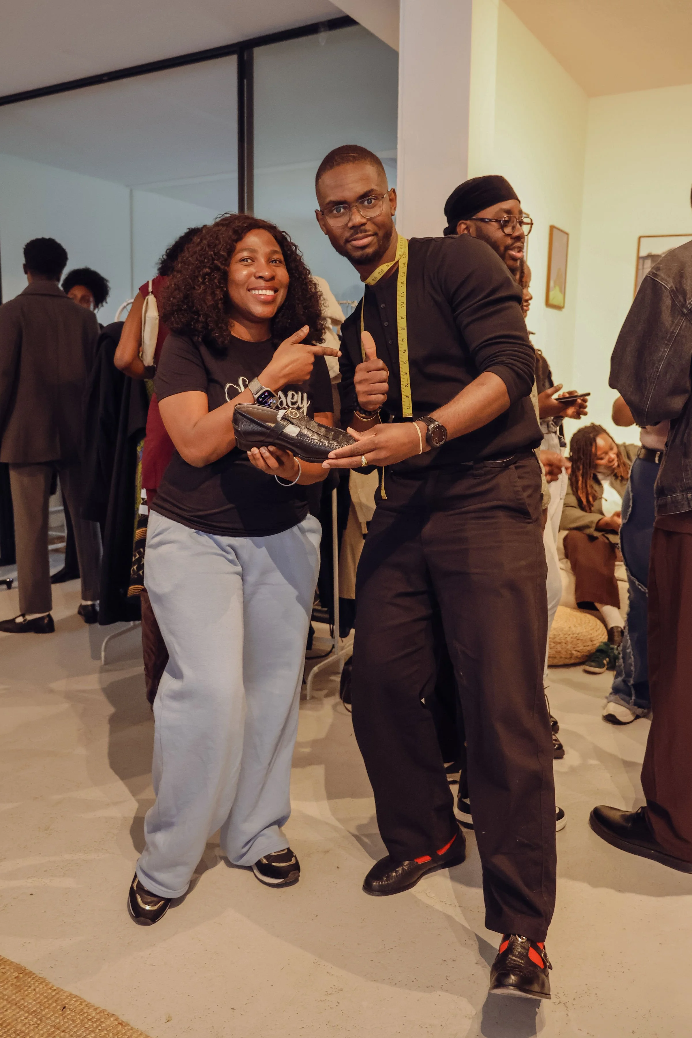 Group of people at an indoor event, with two individuals in the foreground smiling and posing for the camera, one pointing at the other, holding a shoe, and wearing casual clothing, surrounded by others engaged in conversation and activities.