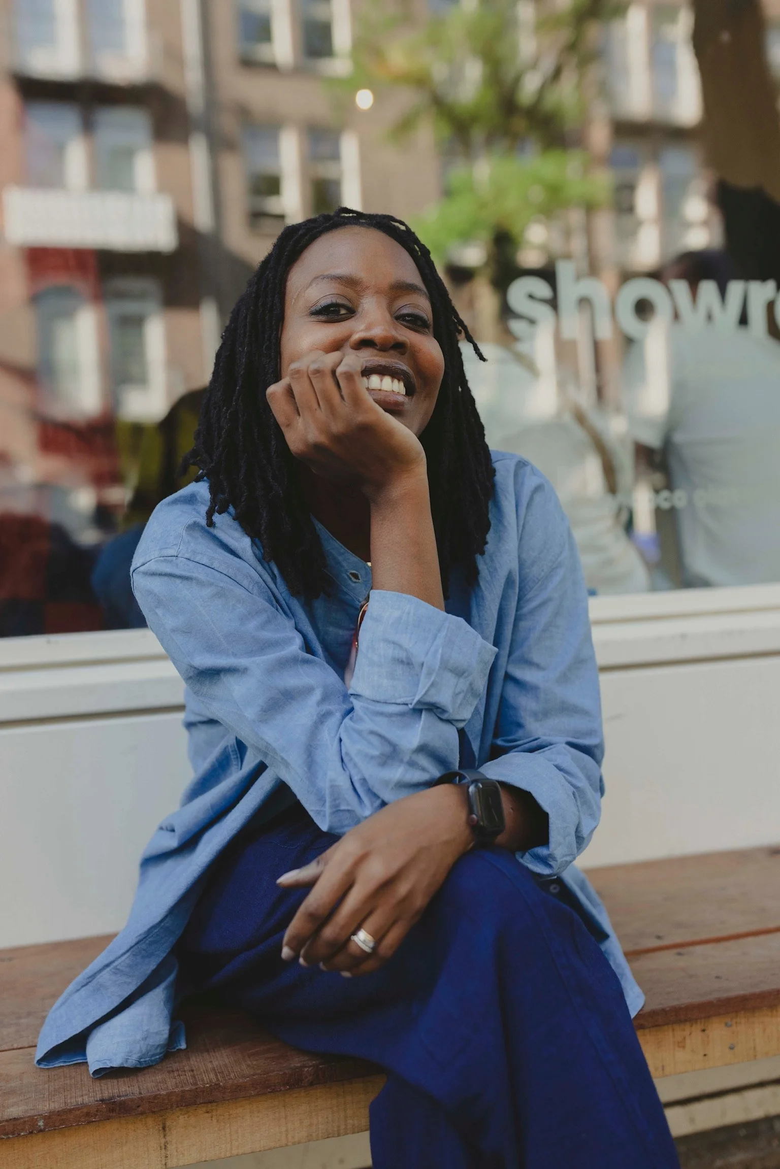 A woman with dark dreadlocks and a blue denim shirt smiling and sitting at a wooden table outside, with buildings and trees in the background.