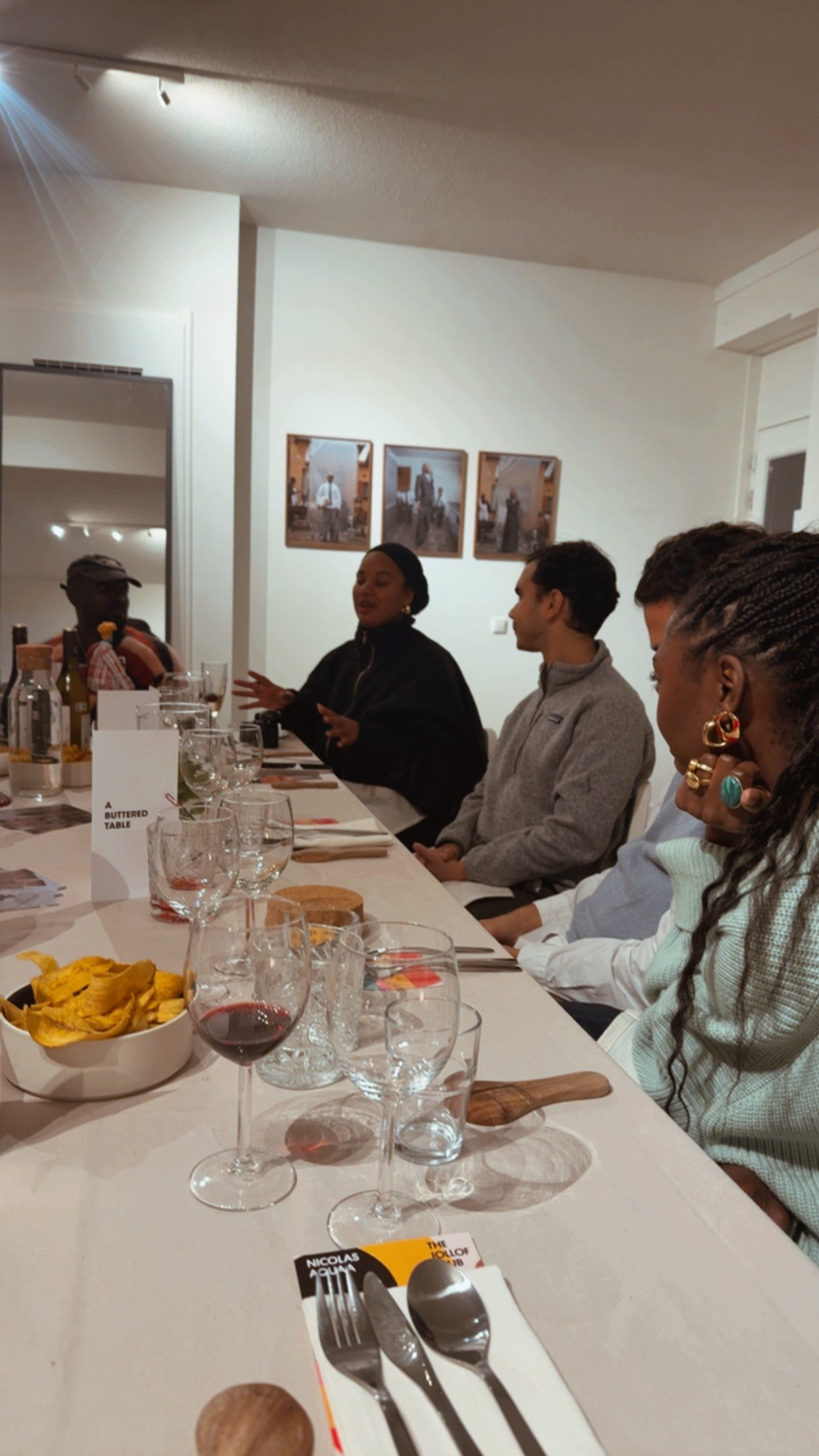 Group of people sitting at a dining table during a dinner event, with plates, glasses, and bowls of snacks, in an indoor setting with art on the wall.