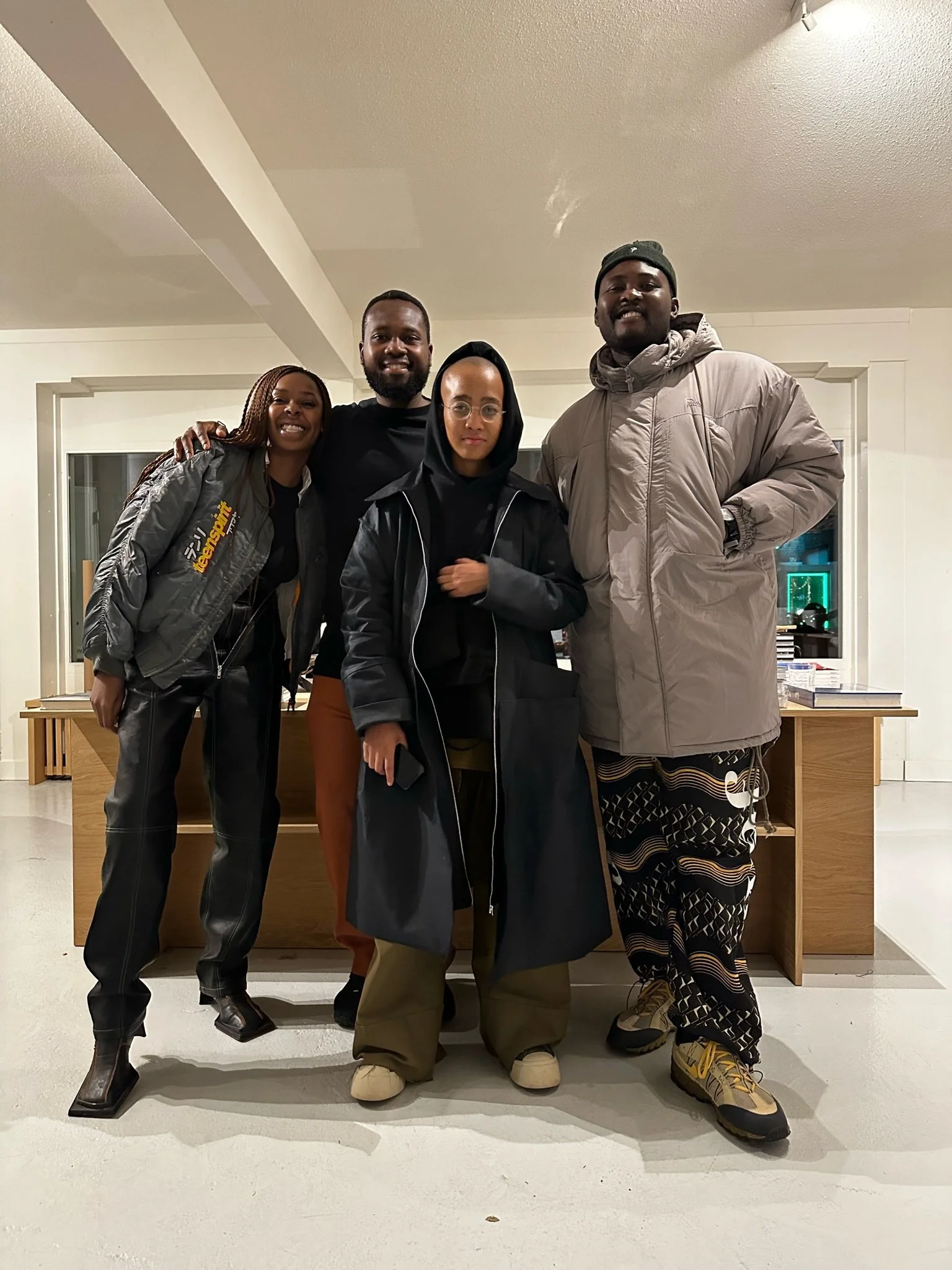 Four people standing together indoors in a room with white walls and a wooden table behind them. They are smiling and dressed in casual and outerwear clothing.