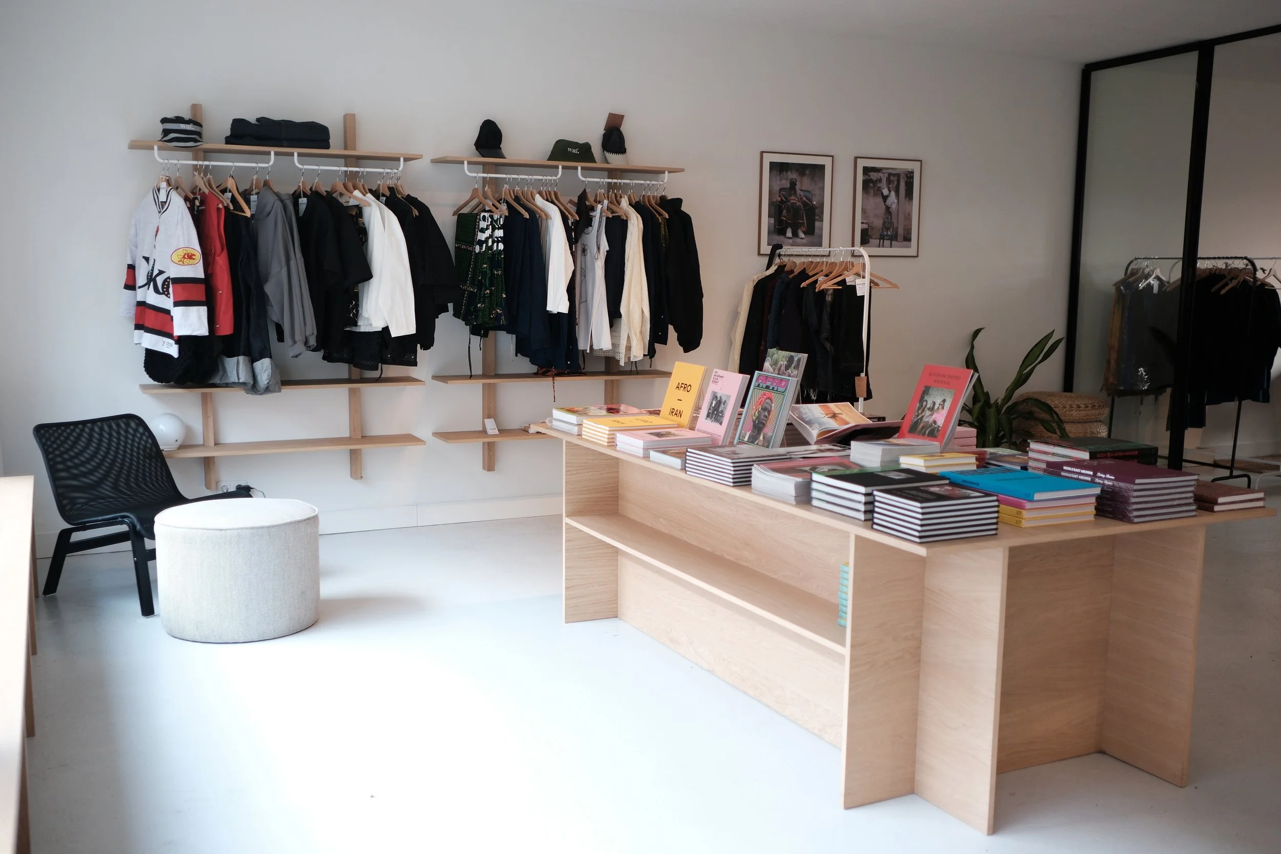 Interior of a boutique clothing store with a white wall and wooden display shelves holding hanging clothes, books, and decorative items, a black chair, a white round stool, and framed artwork on the wall.