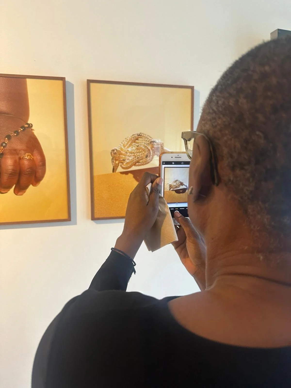 A person taking a photo of two art pieces on a white wall, one of a hand with rings and a bracelet, and the other of a snake, both with a yellow background.