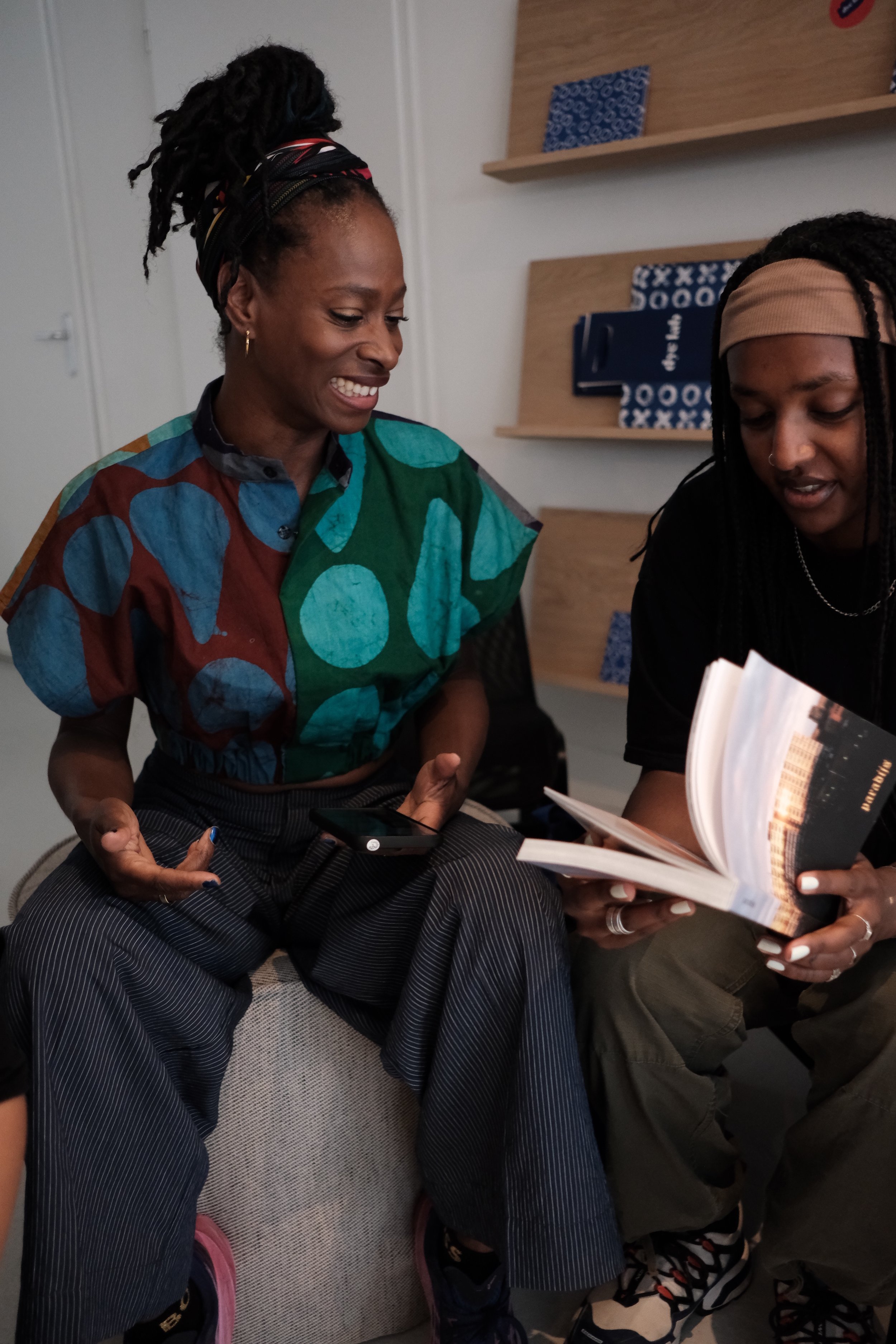 Two women sitting on a gray couch, engaged in conversation. One is holding a book open, and the other is smiling while looking at her phone. The women have dark curly hair, one with dreadlocks and a headband, the other with a colorful patterned top a