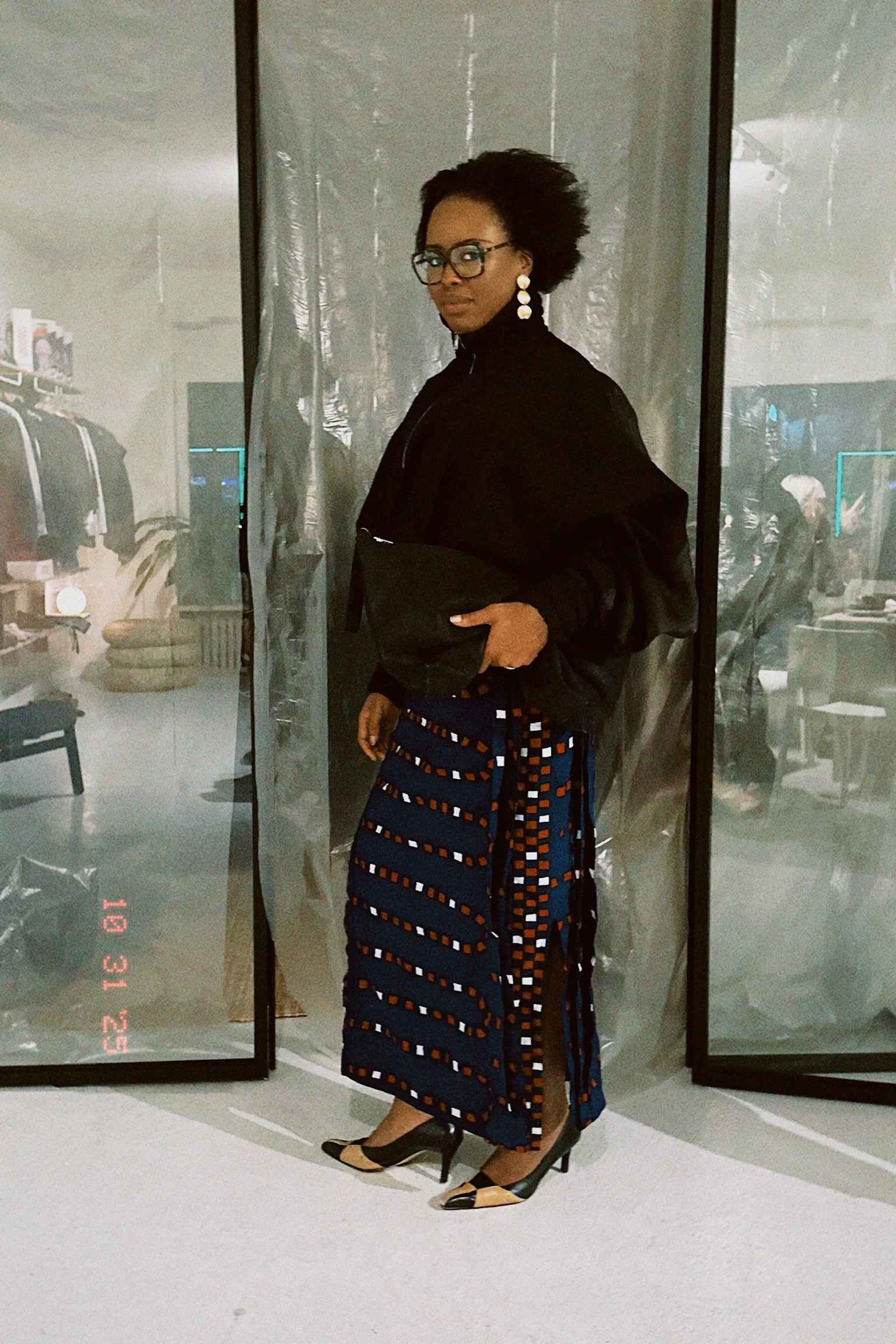 A woman standing indoors, wearing glasses, large white earrings, a black jacket, and a patterned skirt with blue, orange, and white geometric designs. She has one hand in her pocket and is looking at the camera, with a reflective surface behind her s