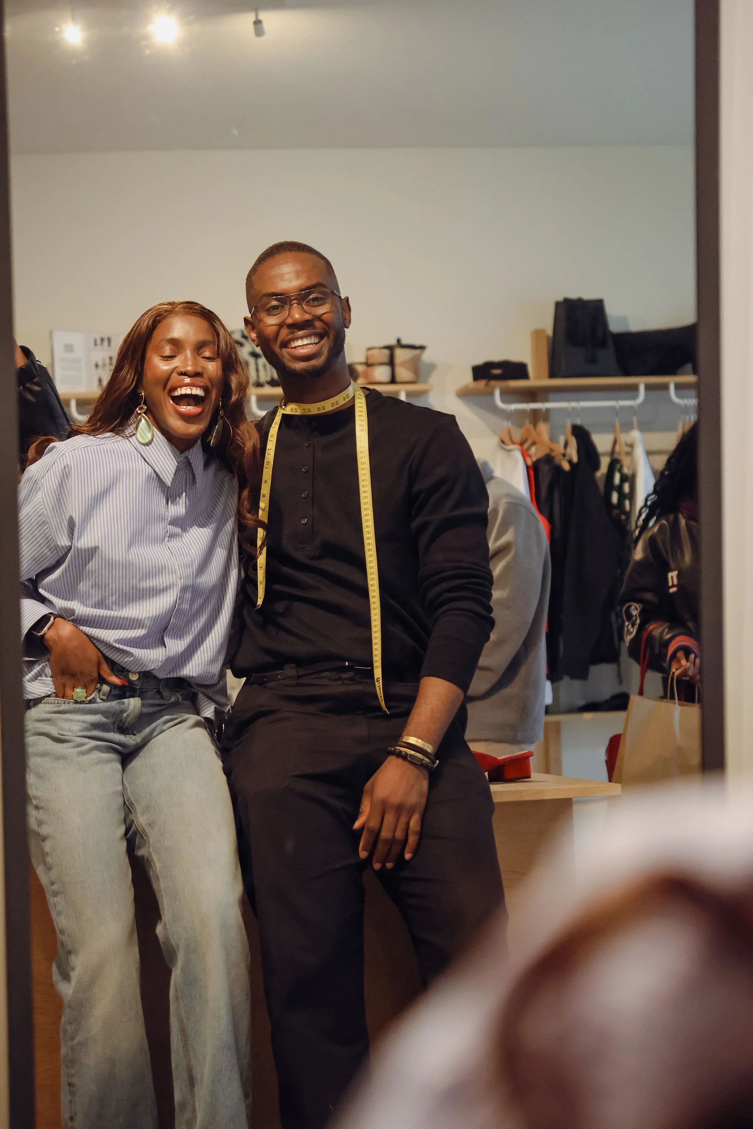 Two young adults, a woman with long wavy hair and earrings and a man with glasses and a measuring tape around his neck, smiling and sitting in a clothing store or boutique with shelves and racks of clothes in the background.