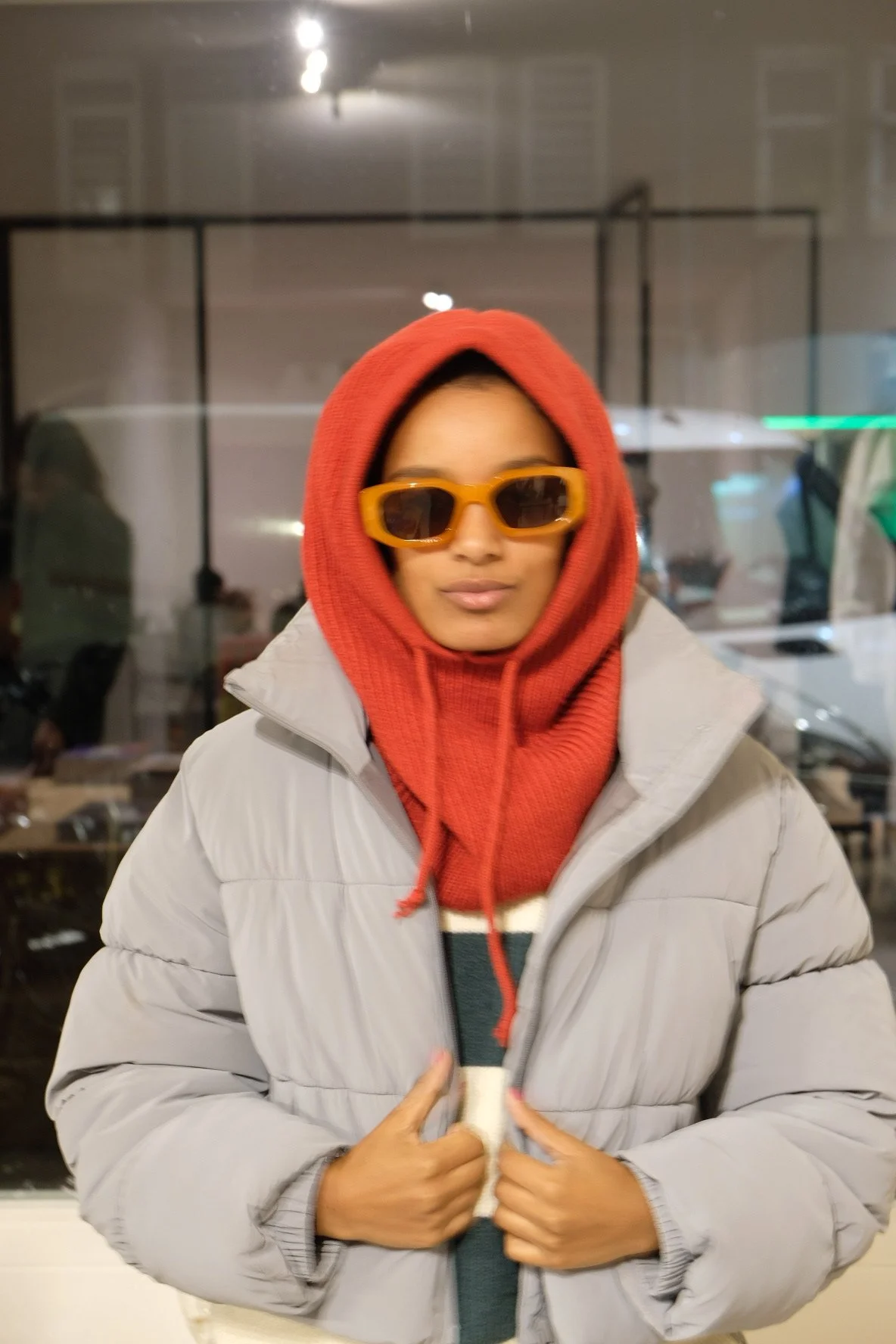 A woman wearing an orange hooded sweater, a light gray puffer jacket, and yellow sunglasses, standing indoors against a glass window with a blurred background.