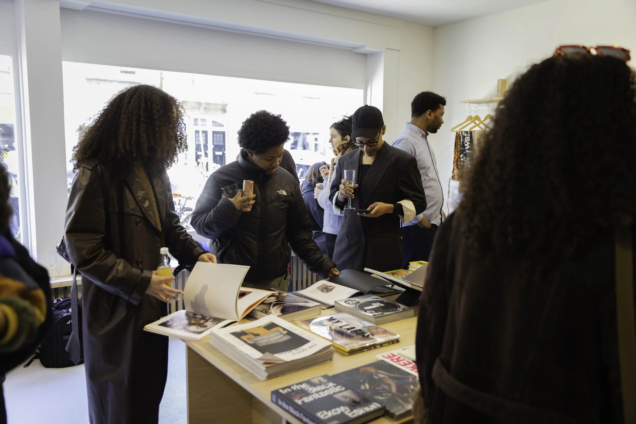 People gathered around a table inside a store or event space, looking at books and magazines displayed on the table. Some are holding drinks, and there is a large window letting in natural light.