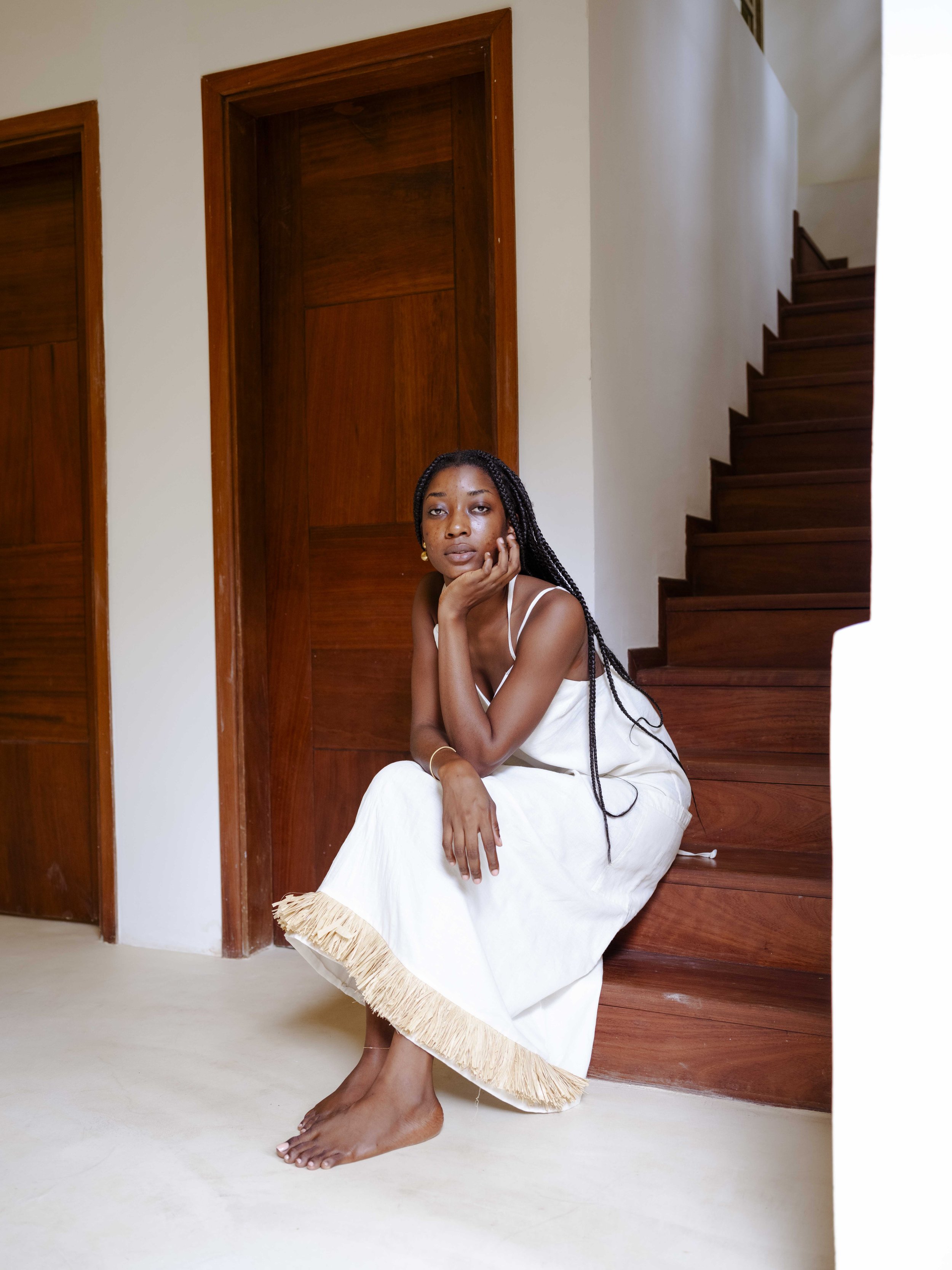 A woman with braided hair and gold earrings sitting on wooden stairs in a minimalist room with wooden door frames and white walls, resting her chin on her hand and looking at the camera.