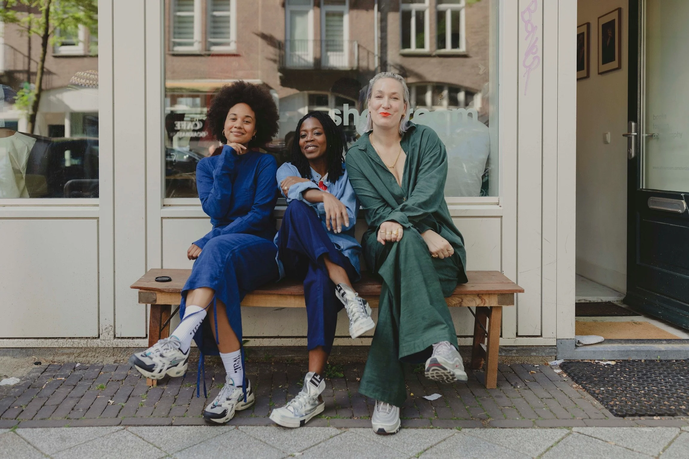 Three women sitting on a wooden bench outside a storefront with large windows. The woman on the left has curly hair and is wearing a blue jacket, the woman in the middle has braided hair and is wearing a blue shirt, and the woman on the right has gra