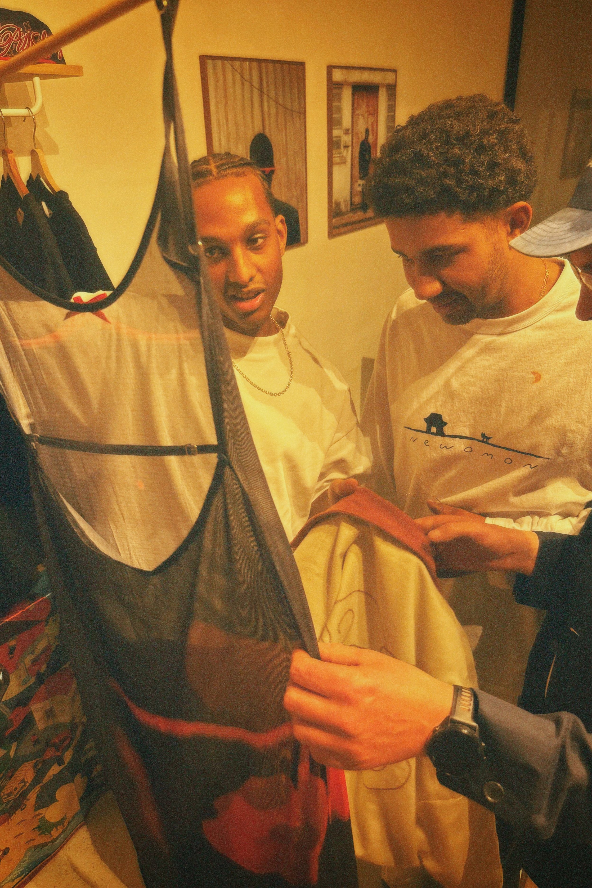 Two young men are seen close together looking down at the person's arm in front of them. The image appears to be taken in a home or casual setting, with artwork on the wall in the background. The left person has braided hair and is wearing a white sh
