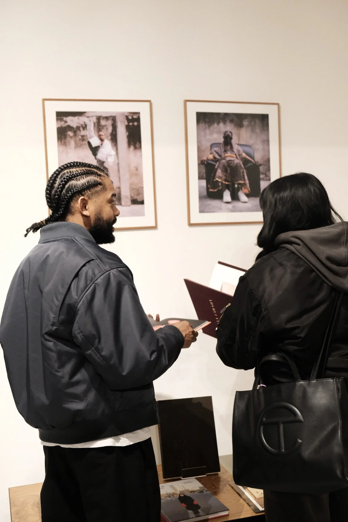 Two people inside an art gallery or museum looking at framed photographs on the wall. One person is holding a booklet or brochure and appears to be explaining or discussing the artwork with the other person.