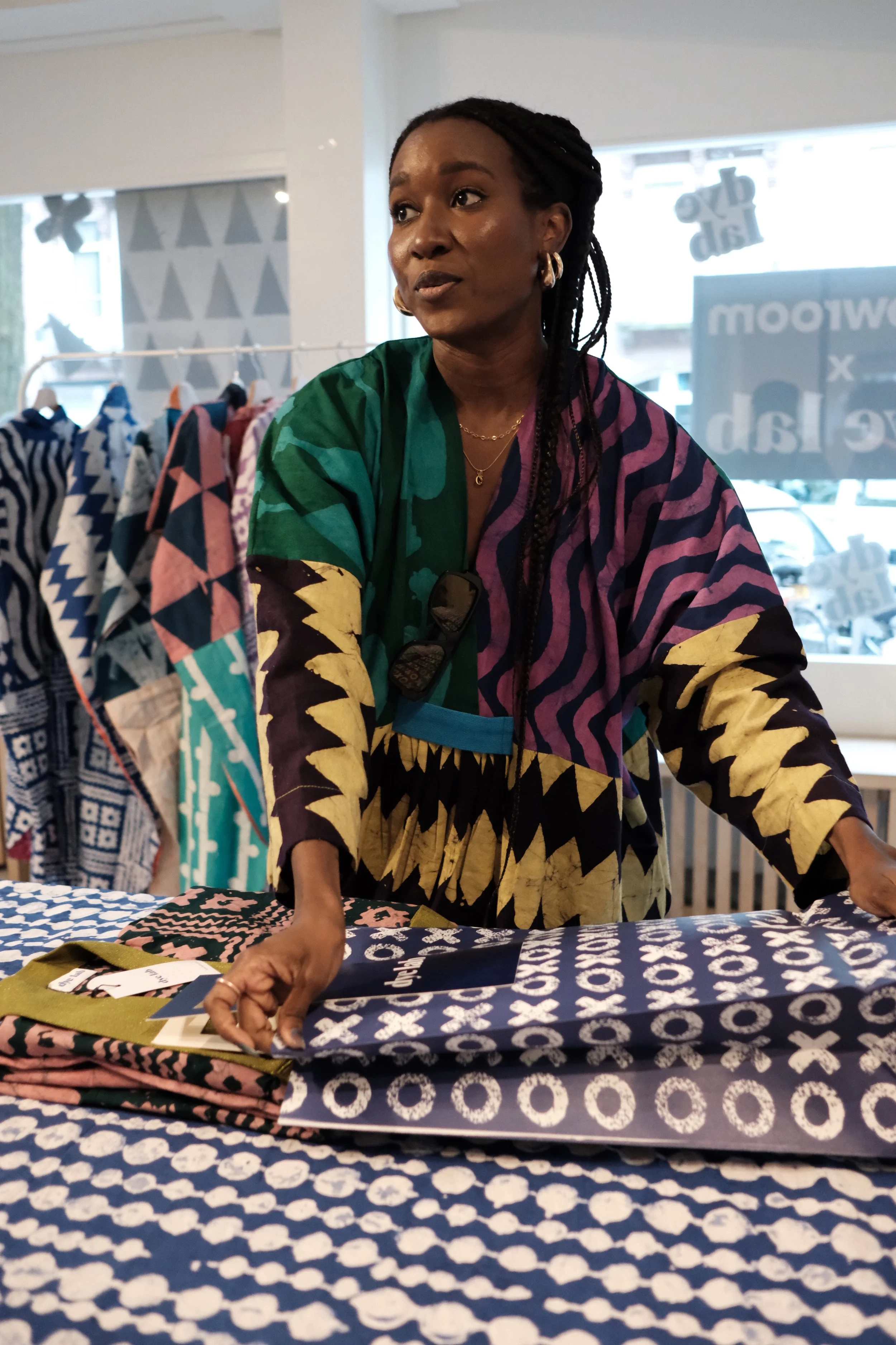 A woman with braided hair, wearing a colorful patterned shirt, stands behind a table displaying various fabric samples in a store.