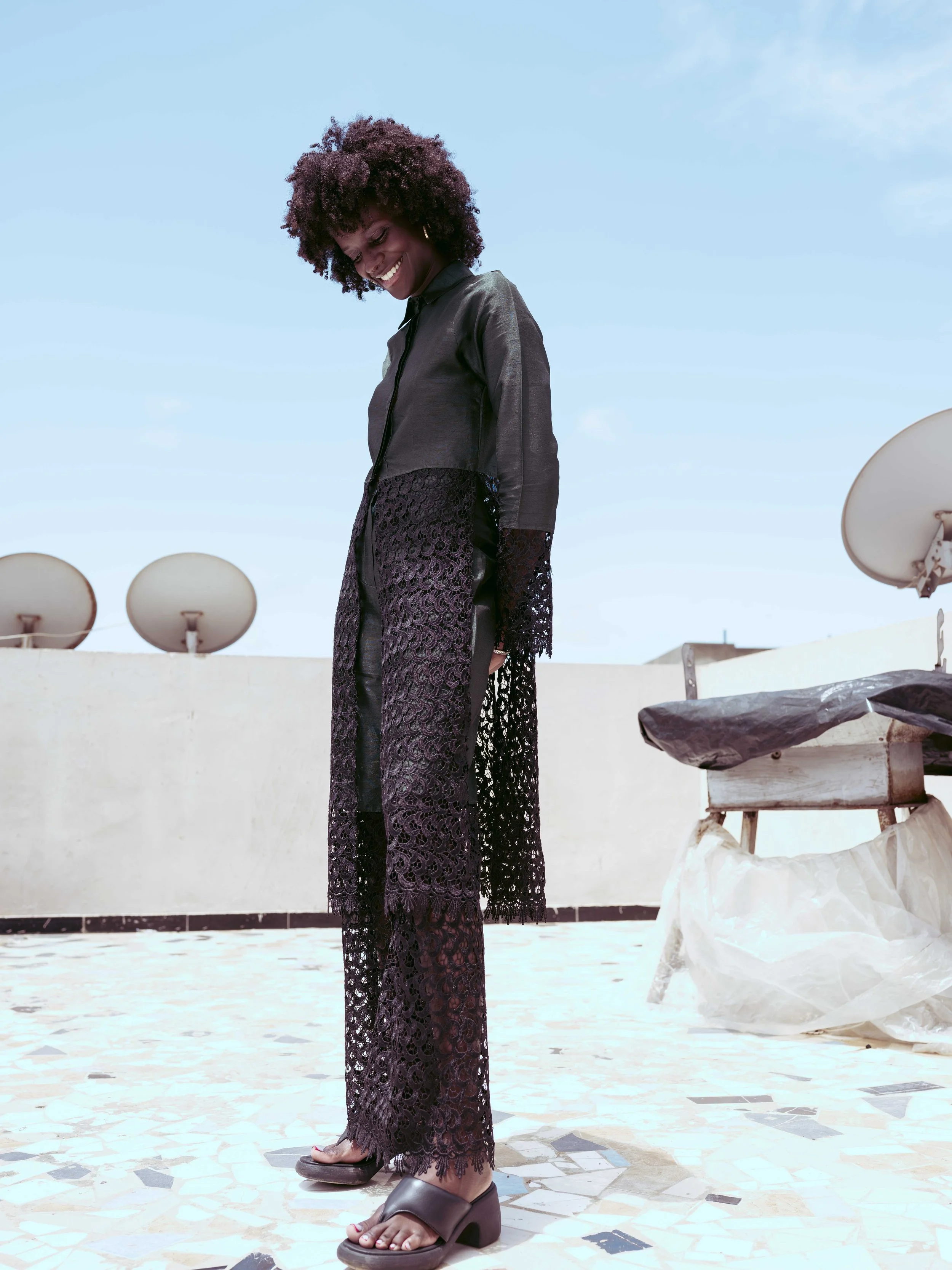 A woman with curly hair smiling and looking down, dressed in a black jacket and a long lace skirt, standing on a rooftop with satellite dishes and a bench nearby, against a blue sky background.
