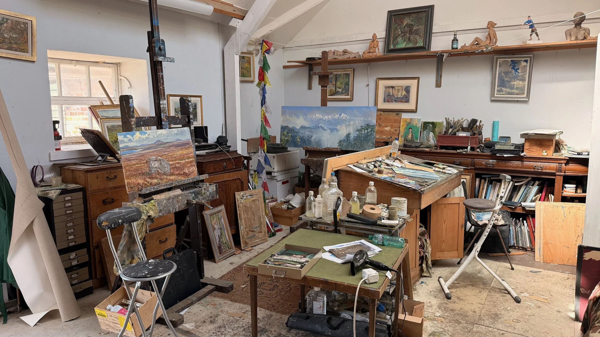 An artist's studio or workshop filled with paintings, art supplies, and furniture. Artwork is displayed on walls and easels, and there are shelves with sculptures and decorative items. The space is cluttered with various tools, bottles, and art piece