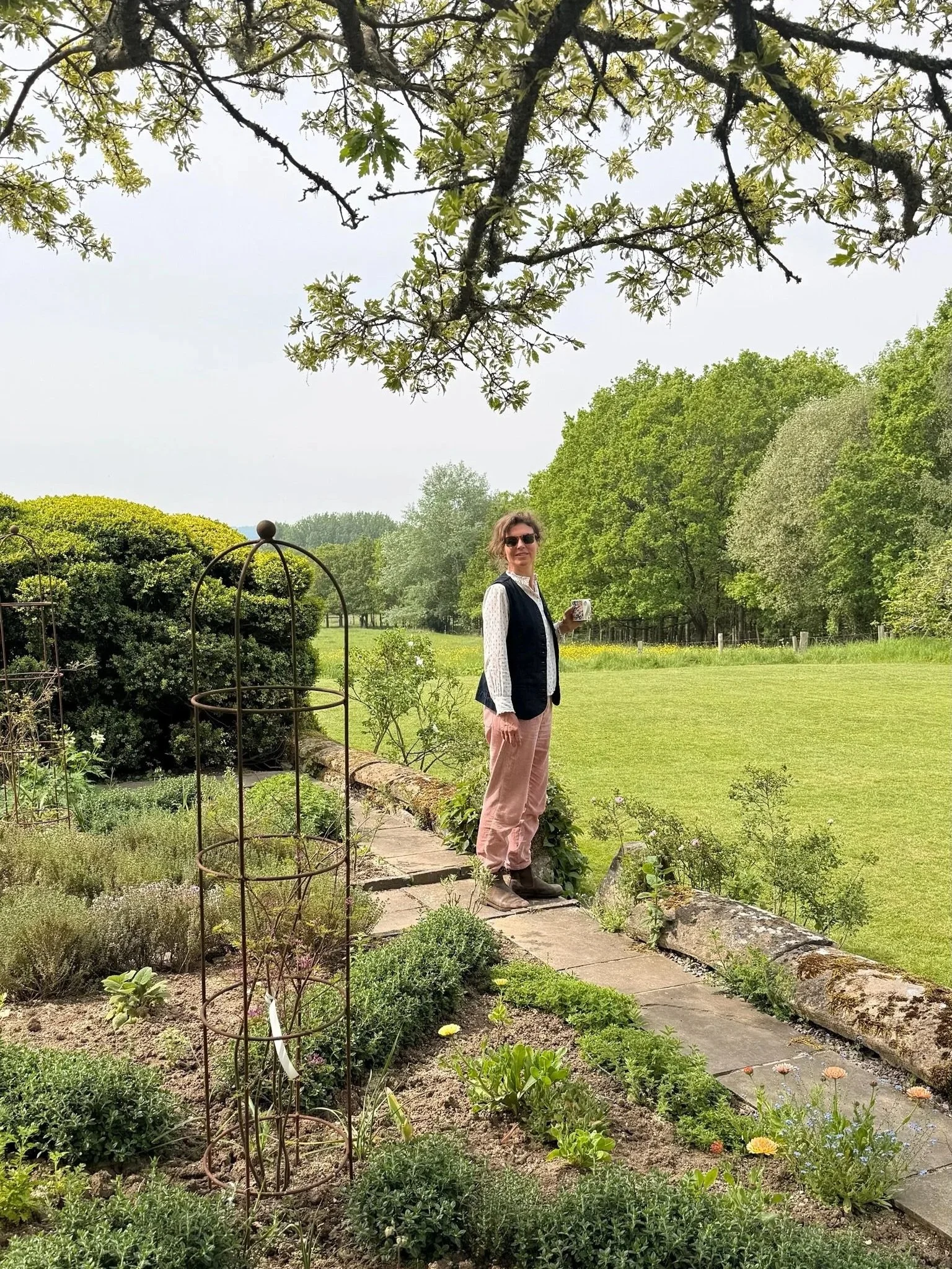 A woman with sunglasses, wearing a black vest, white shirt, pink trousers, and brown boots, stands in a rural garden holding a mug, with trees, bushes and fields in the background.