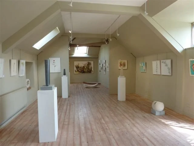 Art gallery in an attic space with artwork on the walls and sculptures on pedestals, wooden floor, and skylight windows.